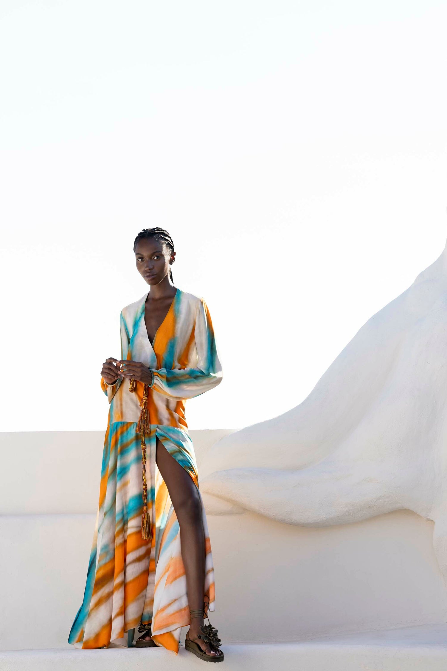 Person wearing a colorful, flowy outfit with a thigh-high slit, standing against a white abstract backdrop.