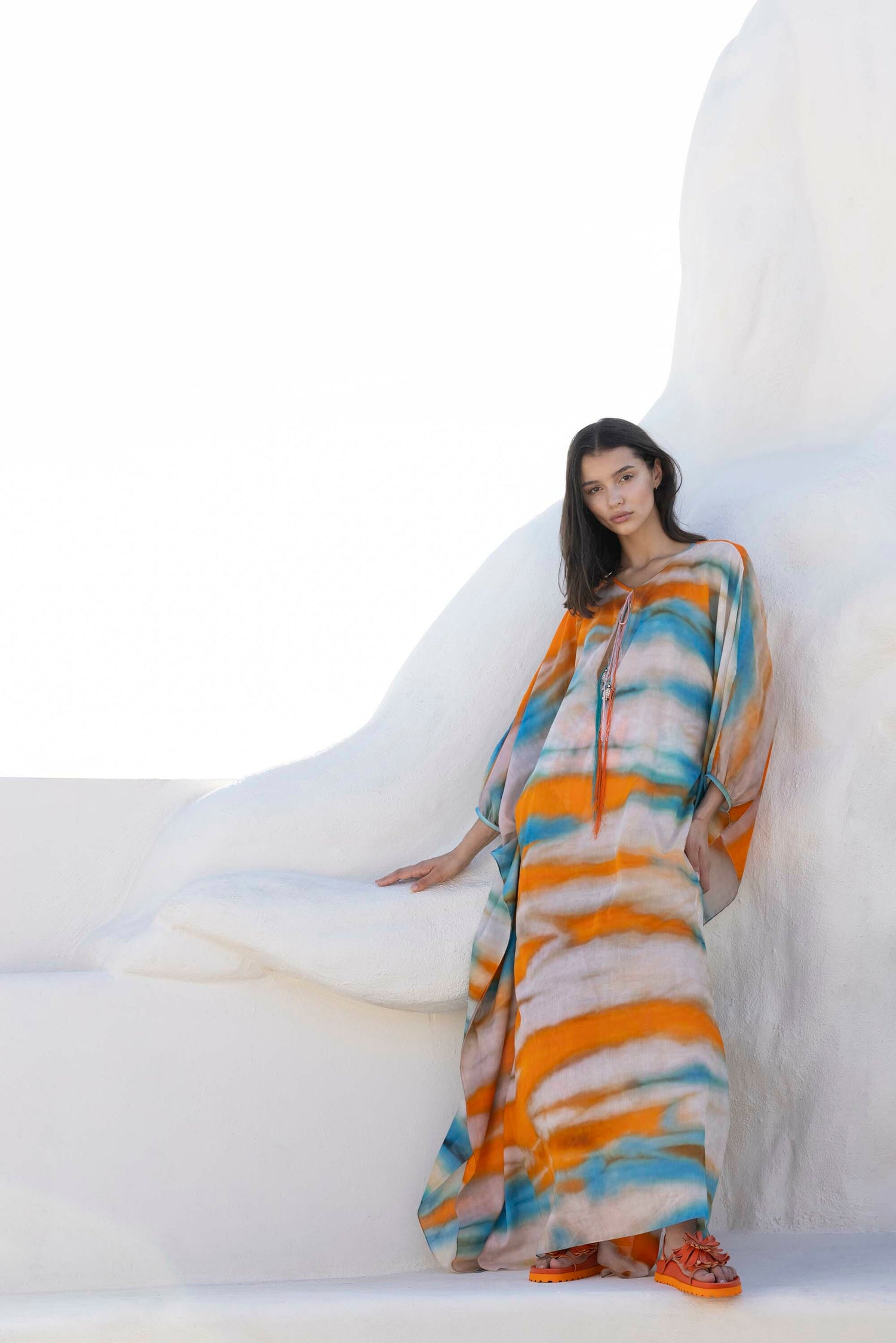 A person in a colorful, flowing dress and orange shoes stands against a white, abstract backdrop.