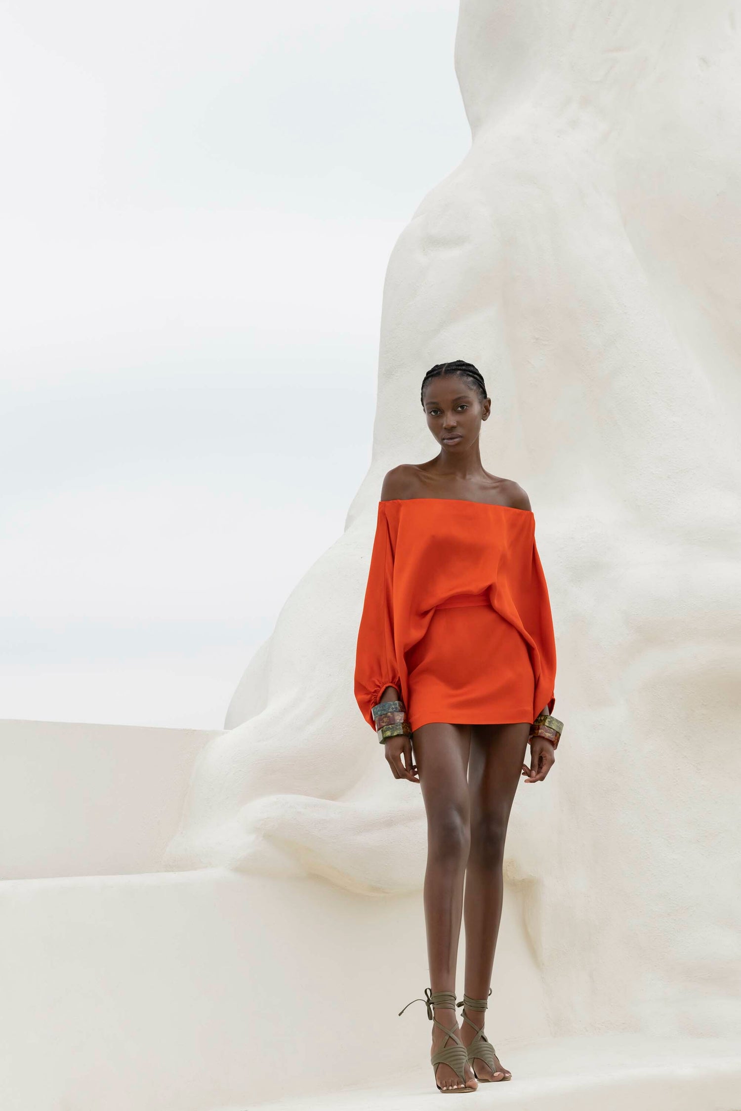 A person in a bright orange off-the-shoulder dress and green sandals stands against a white abstract background.