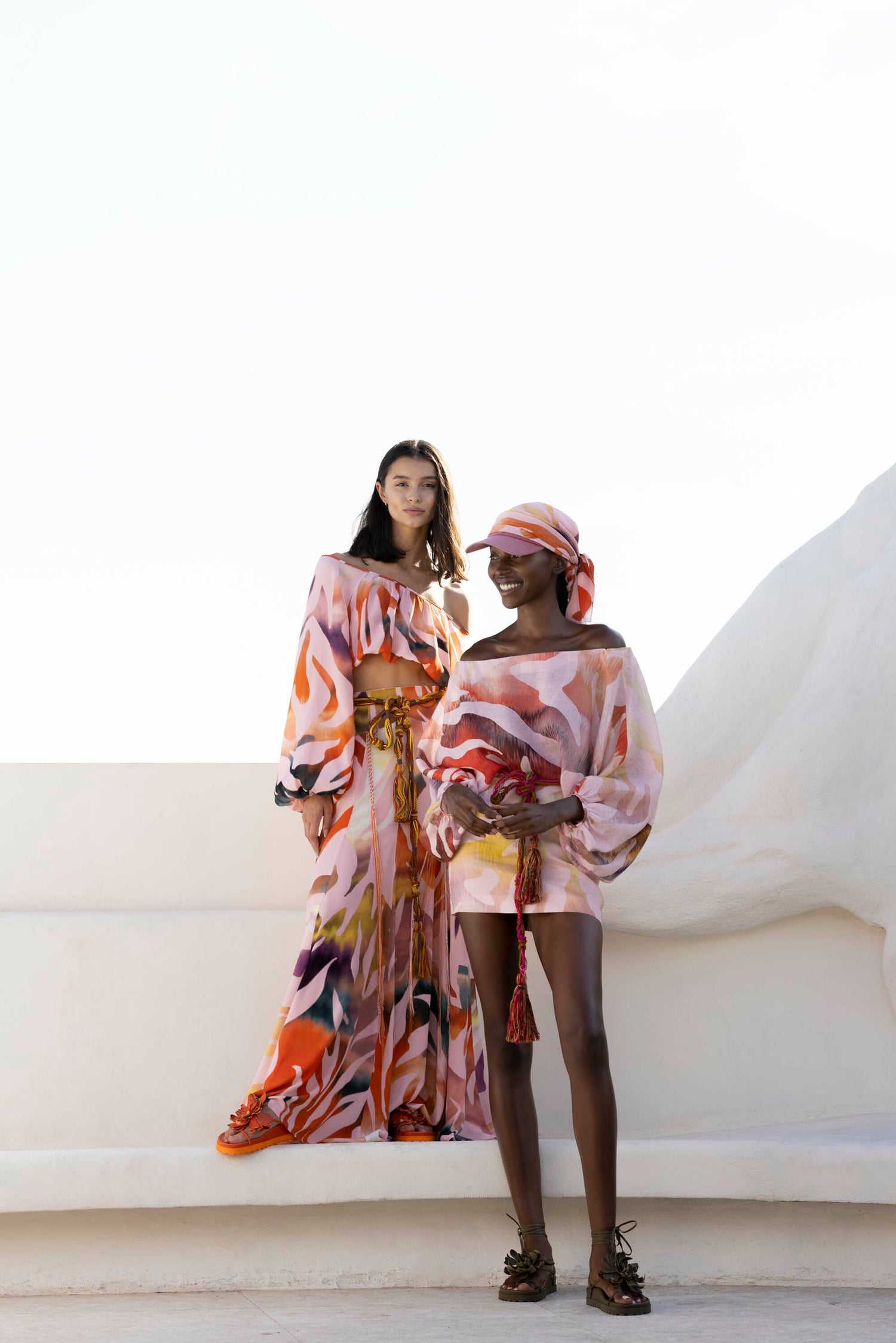 Two women in colorful, flowing outfits pose against a white backdrop.