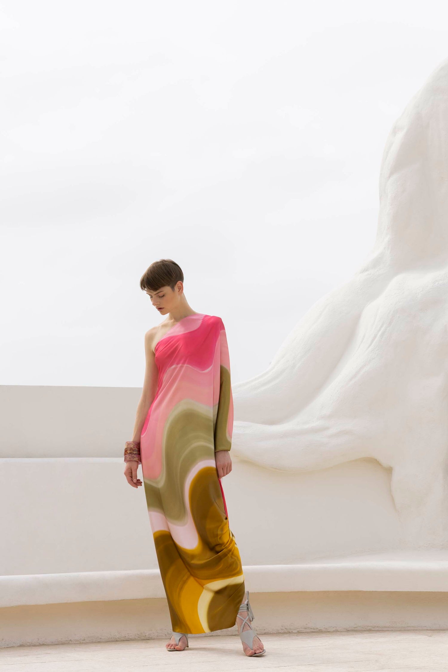 Person in a colorful, flowing dress stands against a white abstract structure, looking down, with a serene expression.