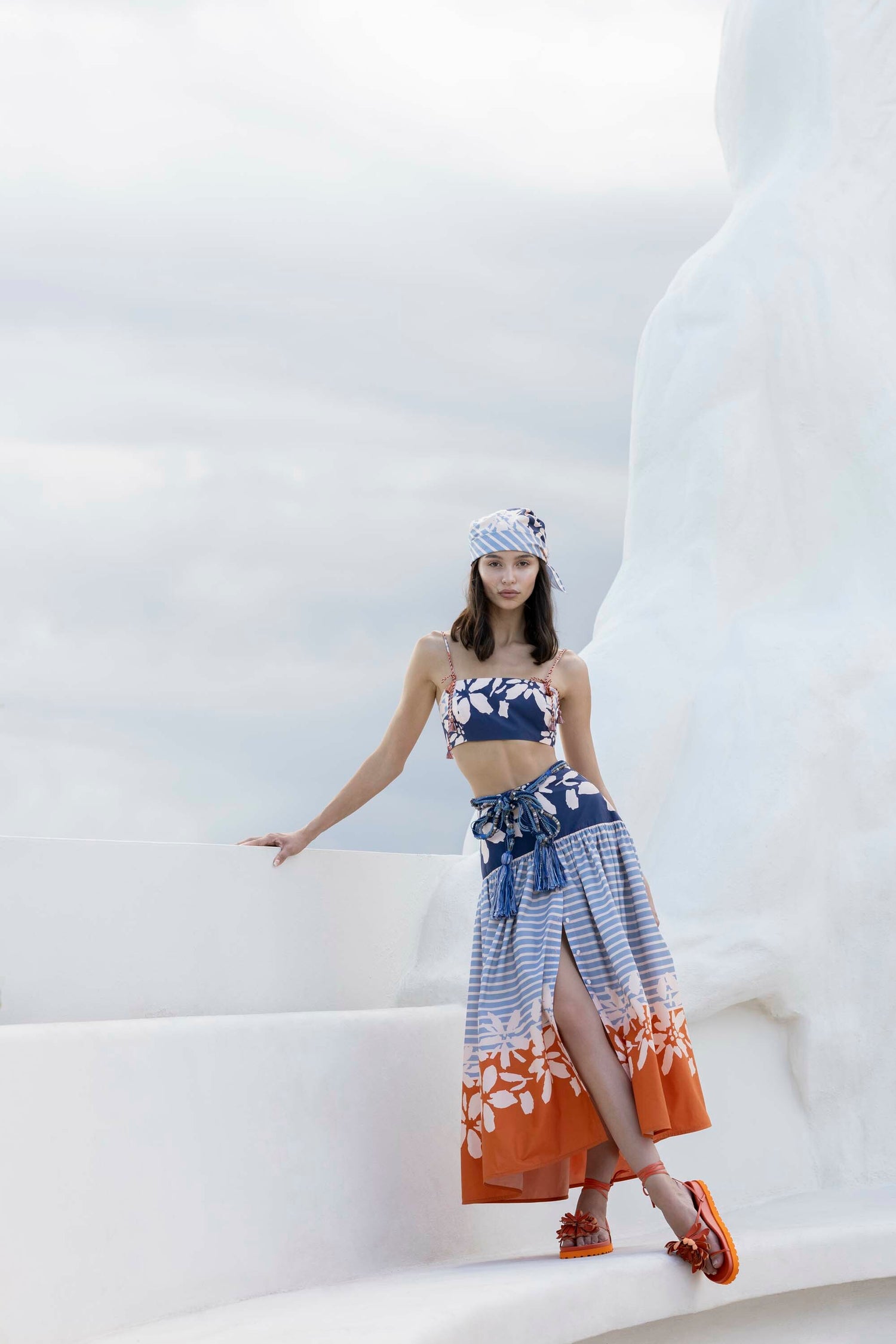 Woman in a blue and orange floral outfit with matching headscarf poses against a white structure under a cloudy sky.