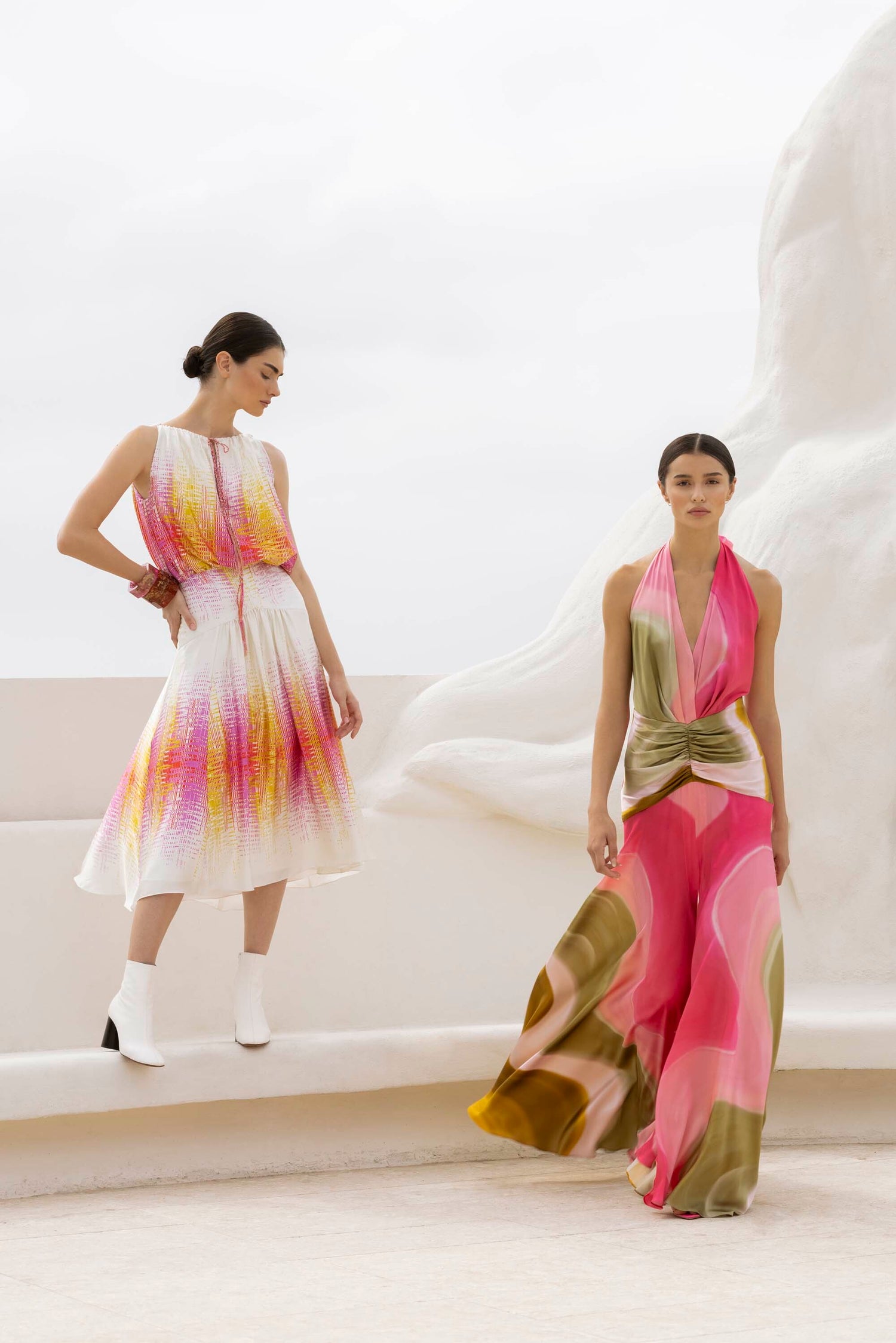 Two women modeling colorful, flowing dresses. One wears a sleeveless dress with pink and yellow patterns, and the other a pink and green halter top and skirt. Both have hair tied back.