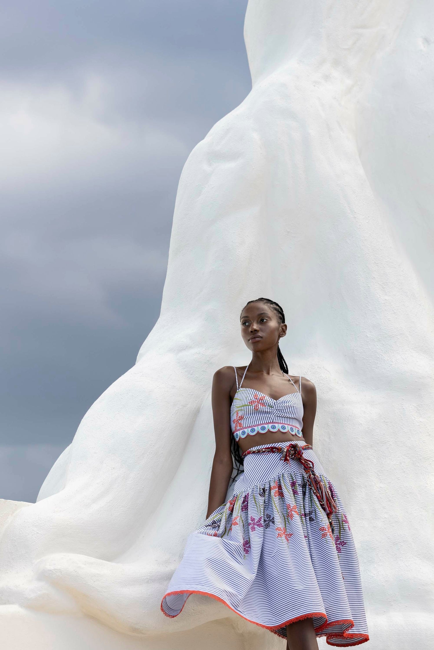 Person wearing a patterned dress leans against a large, white sculpture with a cloudy sky in the background.