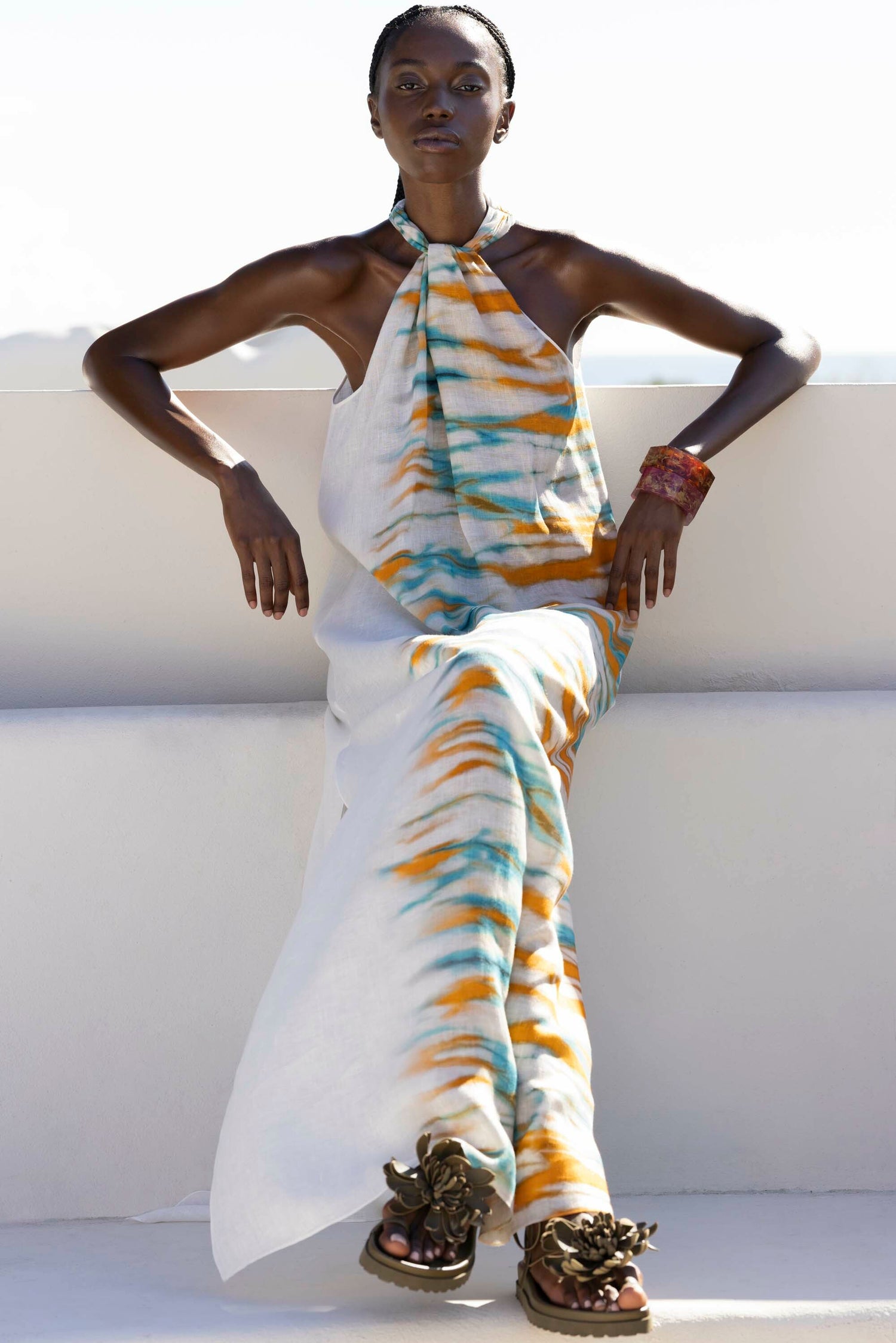 Person sitting in a colorful tie-dye halter dress and sandals against a plain background.