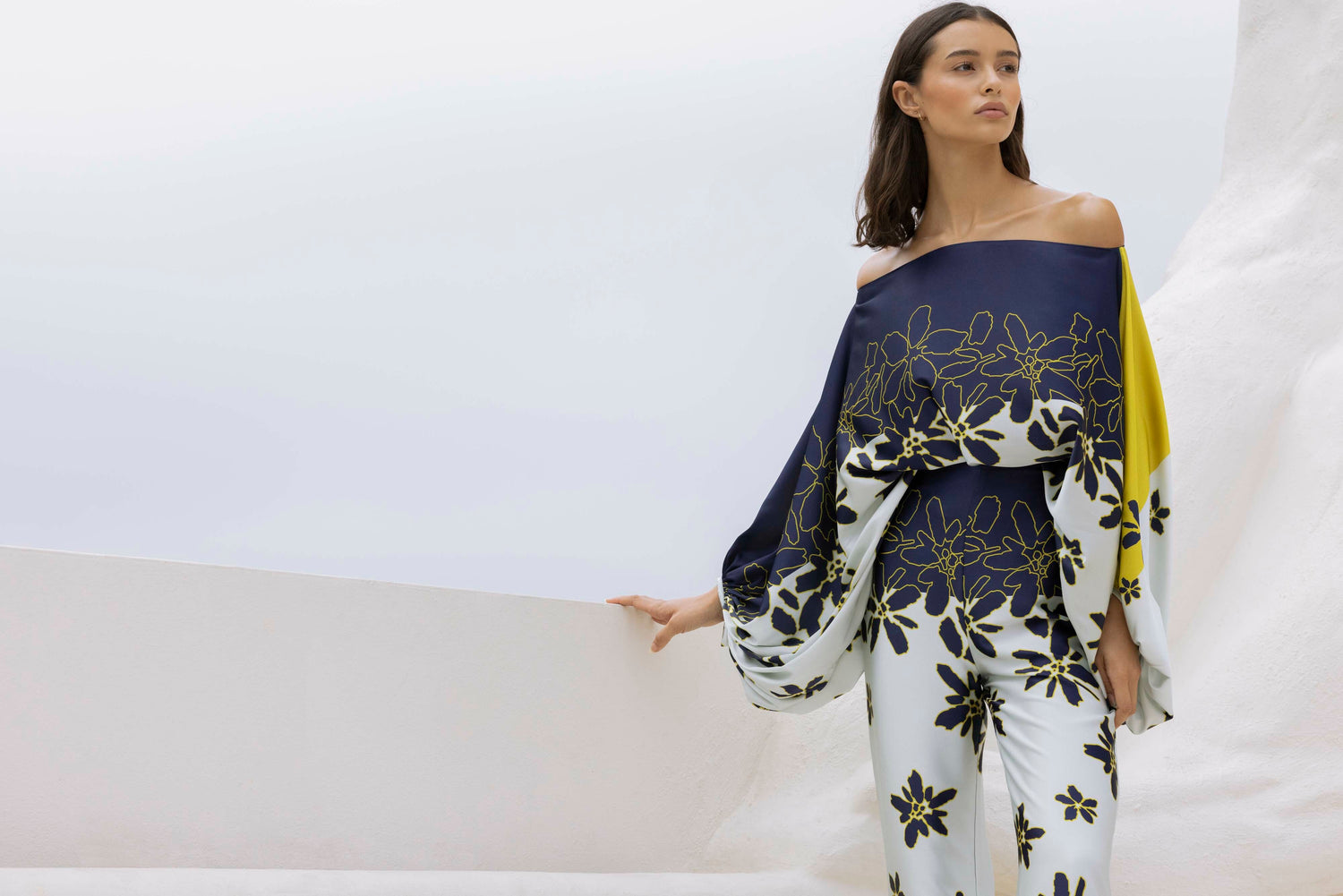 A woman wearing a stylish off-shoulder outfit with a floral pattern stands against a minimalist white background.