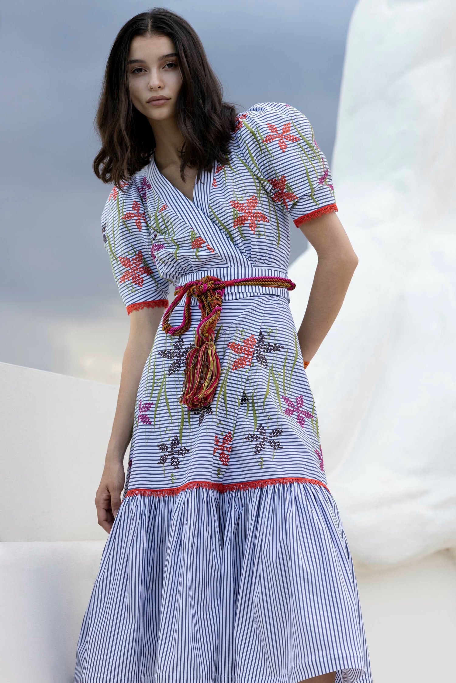 A woman stands outdoors wearing a blue and white striped dress with floral patterns and puffed sleeves, against a cloudy sky backdrop.