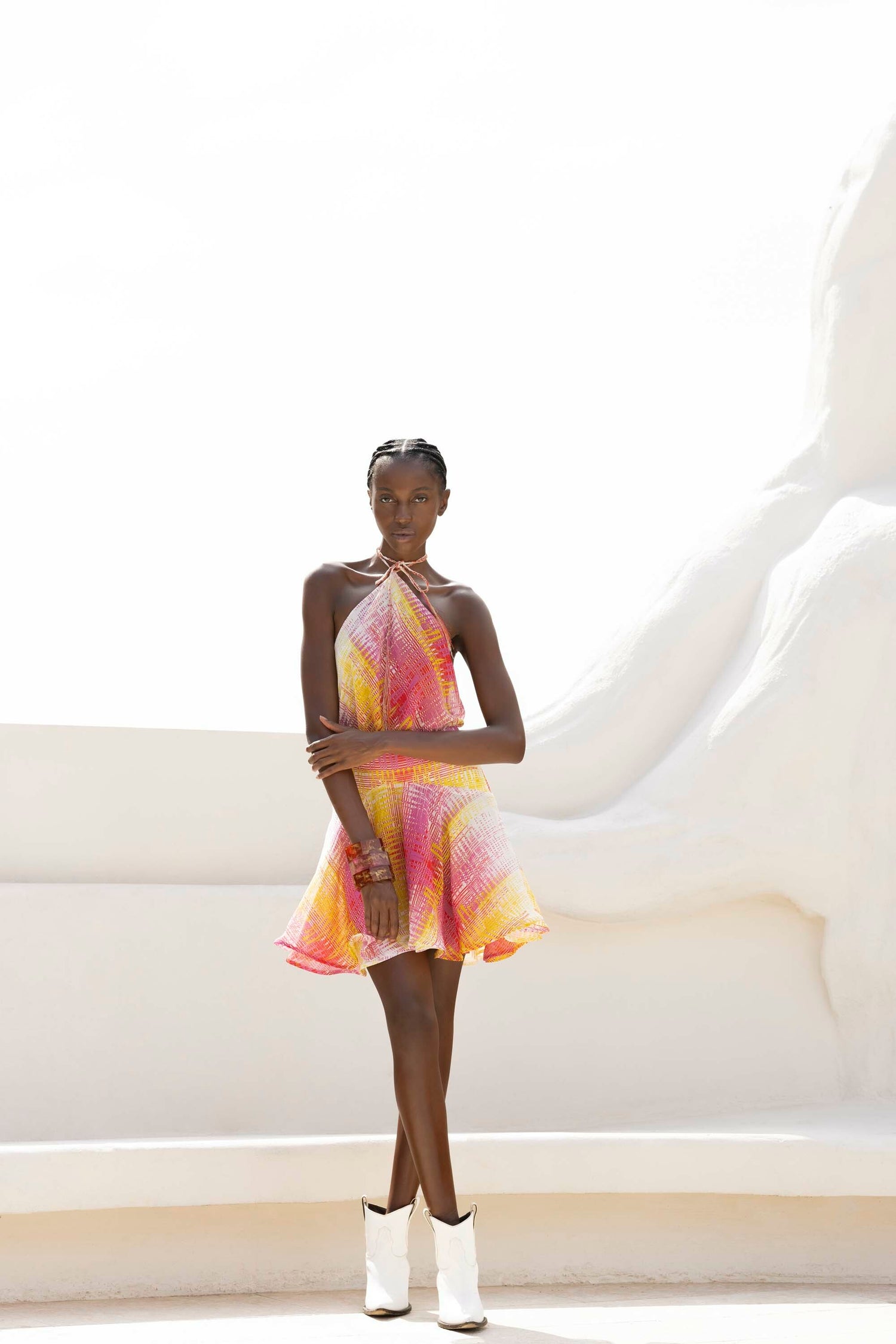 A person stands on a white patio wearing a colorful halter dress and white boots.