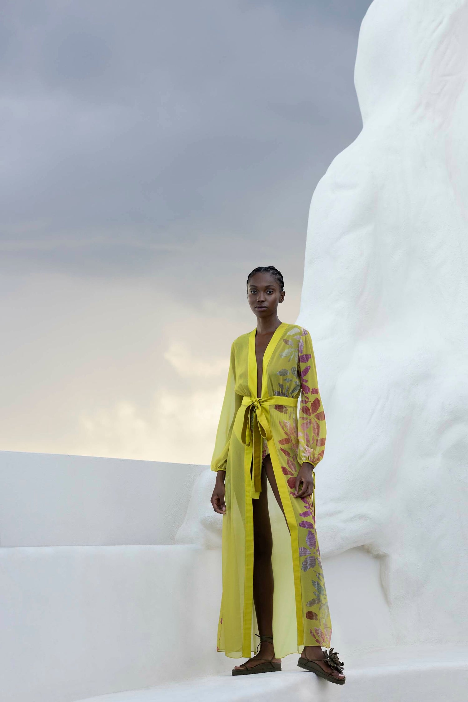 A person stands outdoors wearing a long, sheer yellow dress with floral patterns, against a backdrop of white structures and a gray sky.