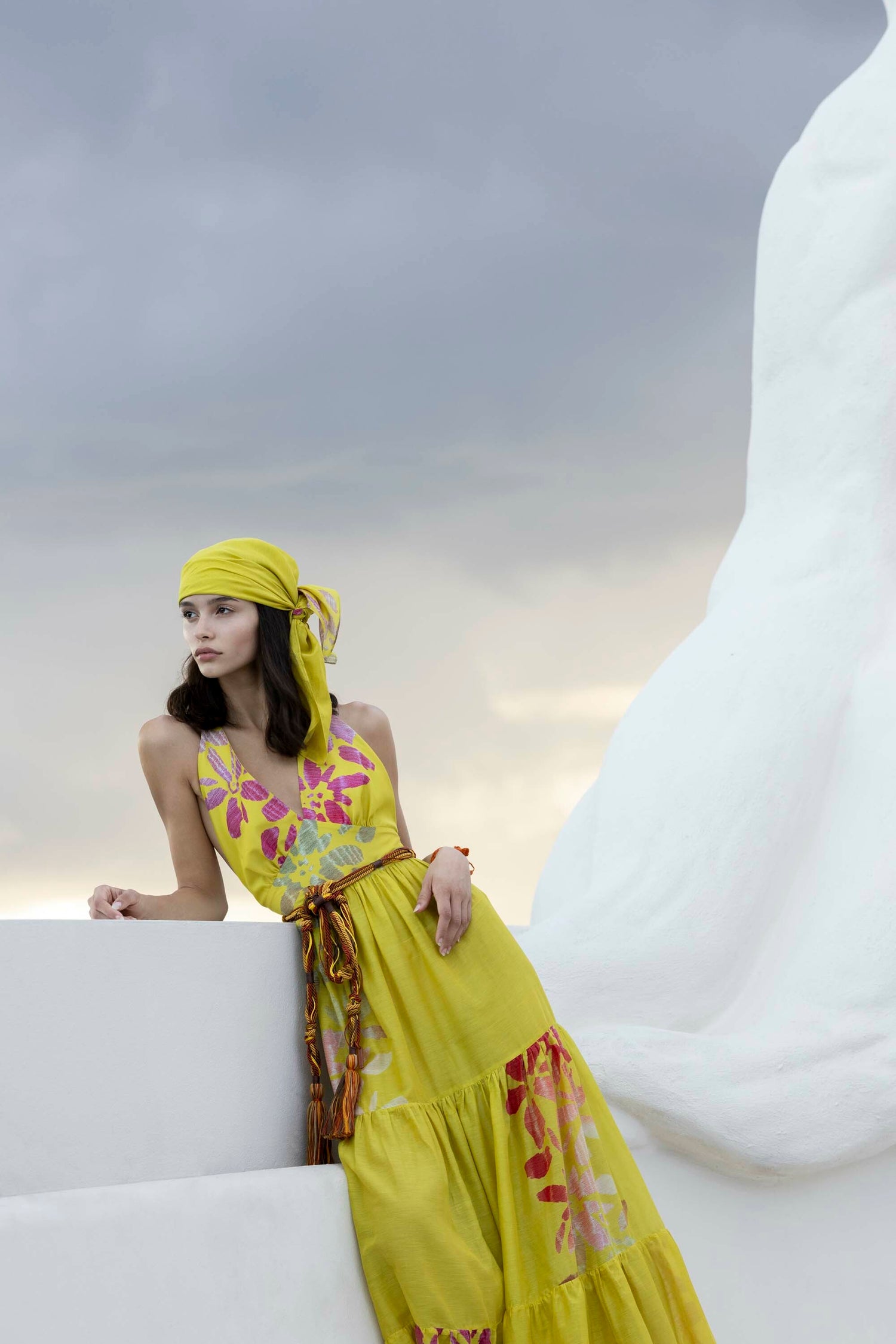 A woman wearing a yellow dress and headscarf leans against a white structure with a cloudy sky in the background.