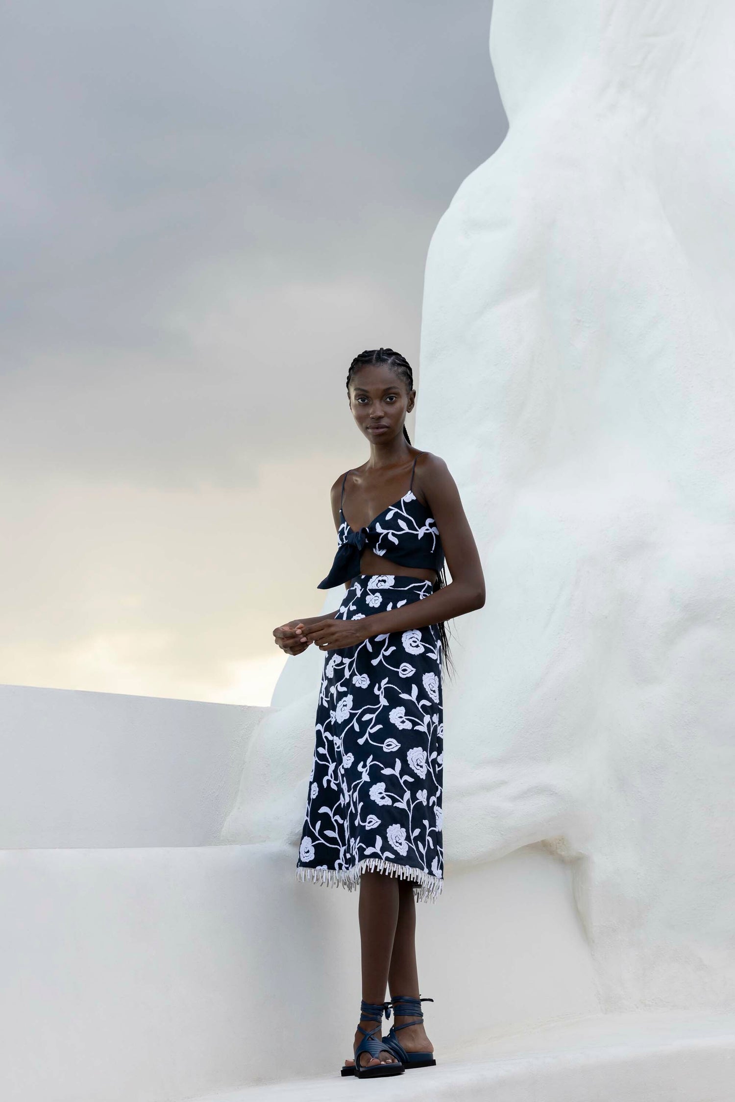 A woman in a black and white floral dress stands outdoors against a light-colored, organic-shaped backdrop under a cloudy sky.