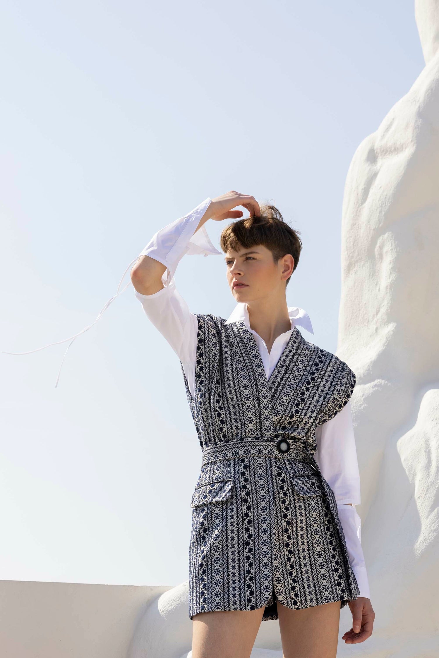 Person in a patterned vest and white shirt poses against a light background, looking to the side with one hand on their head.