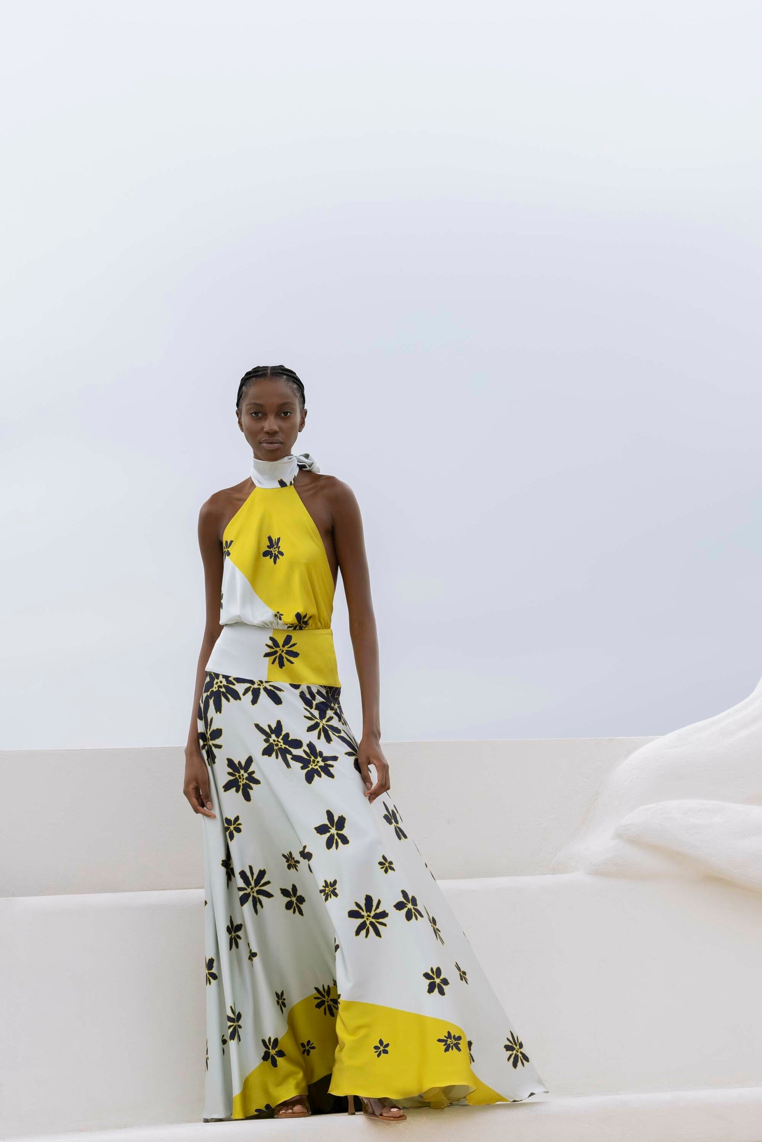 A woman stands against a minimal backdrop wearing a sleeveless dress with a yellow and white floral pattern.