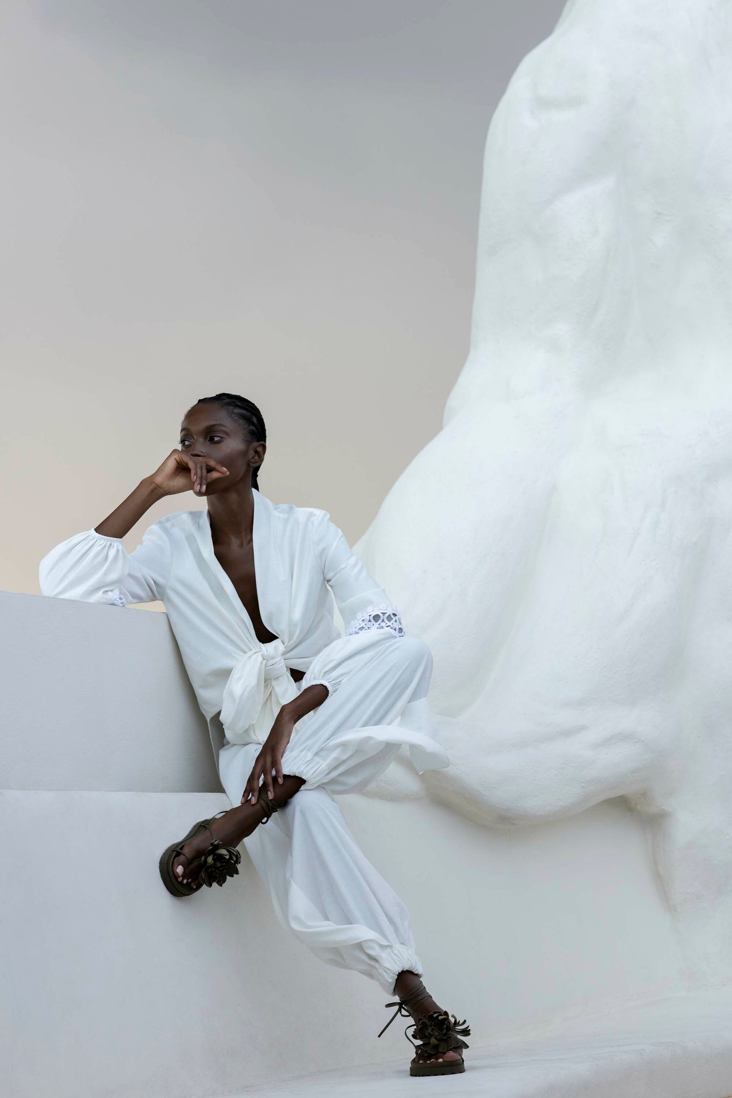 A person in a white outfit sits pensively on a white structure, one arm resting on their knee. A large abstract white sculpture fills the background.