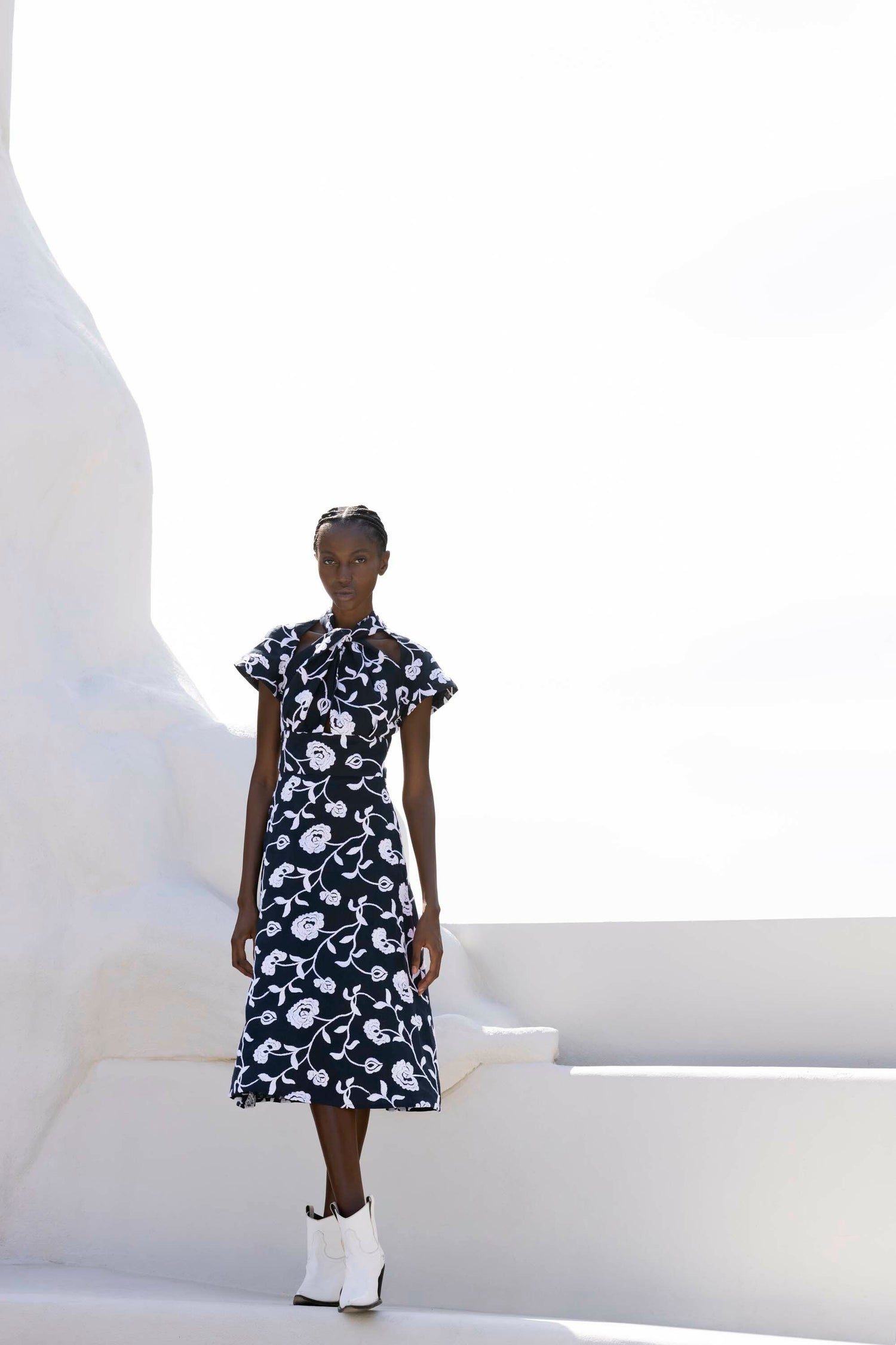 A person in a floral black and white dress and white boots stands against a minimalist white background.