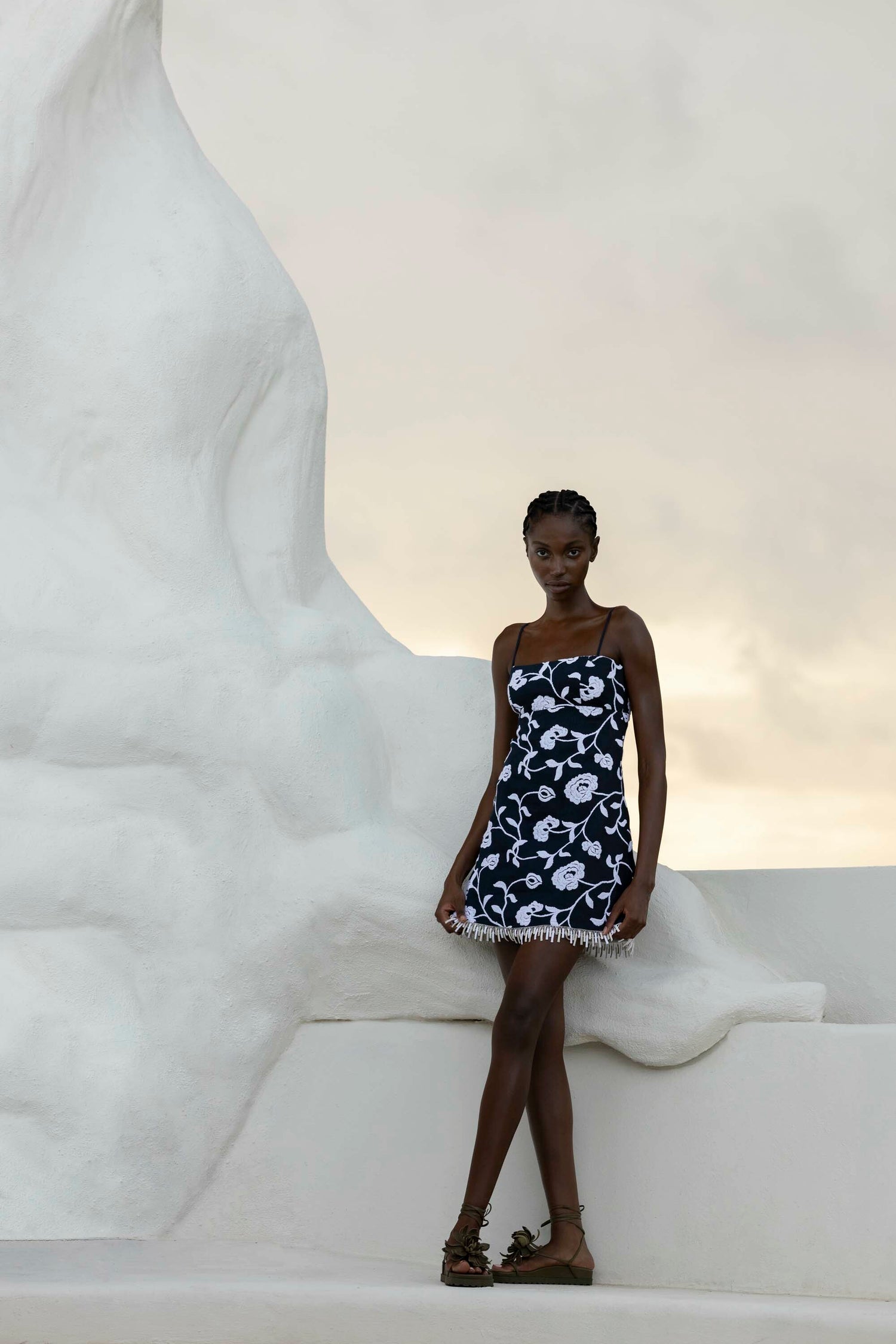 Person wearing a patterned dress stands against a large white sculpture with a cloudy sky backdrop.