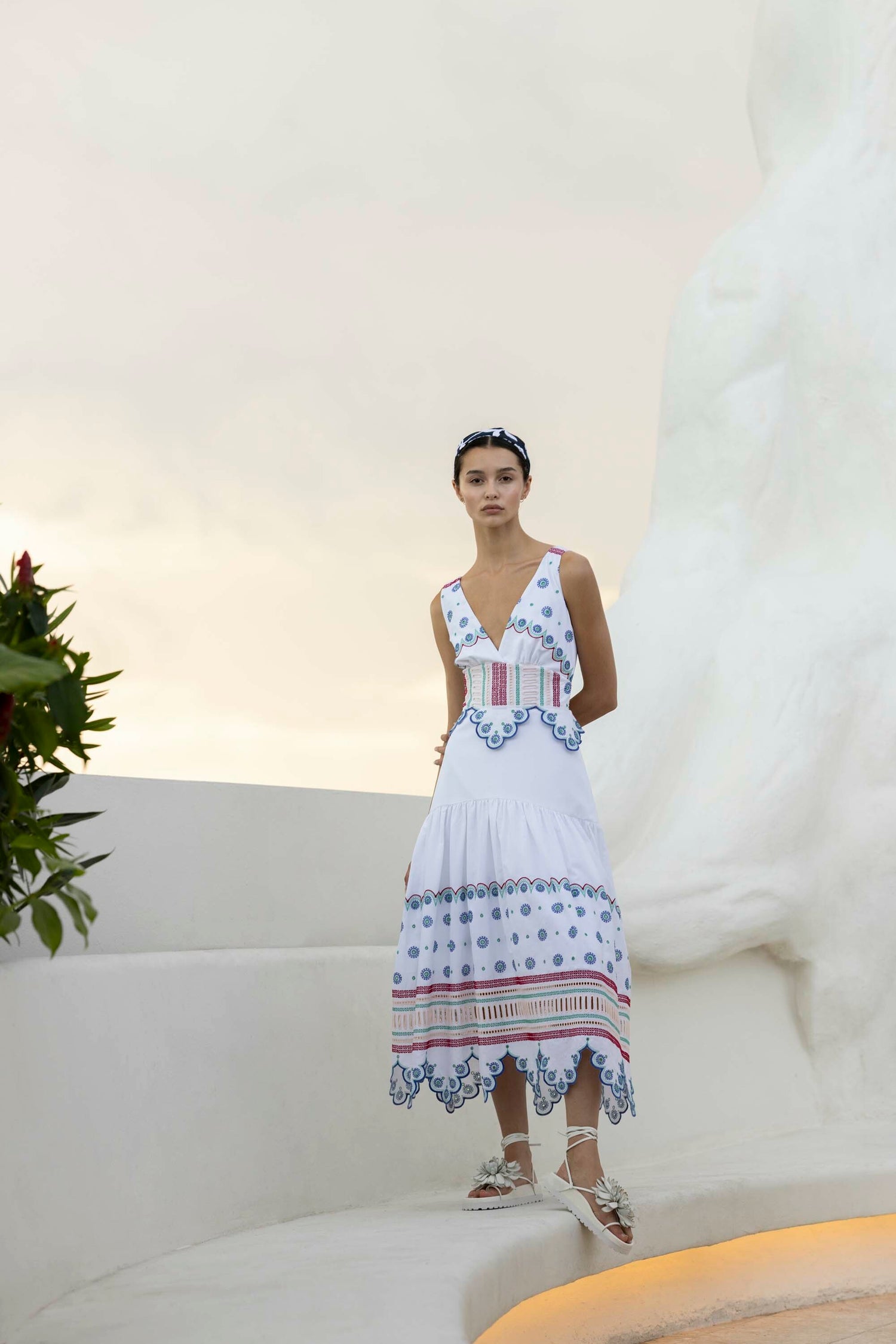 A woman in a white and blue patterned dress stands beside a large white statue with greenery nearby under a cloudy sky.