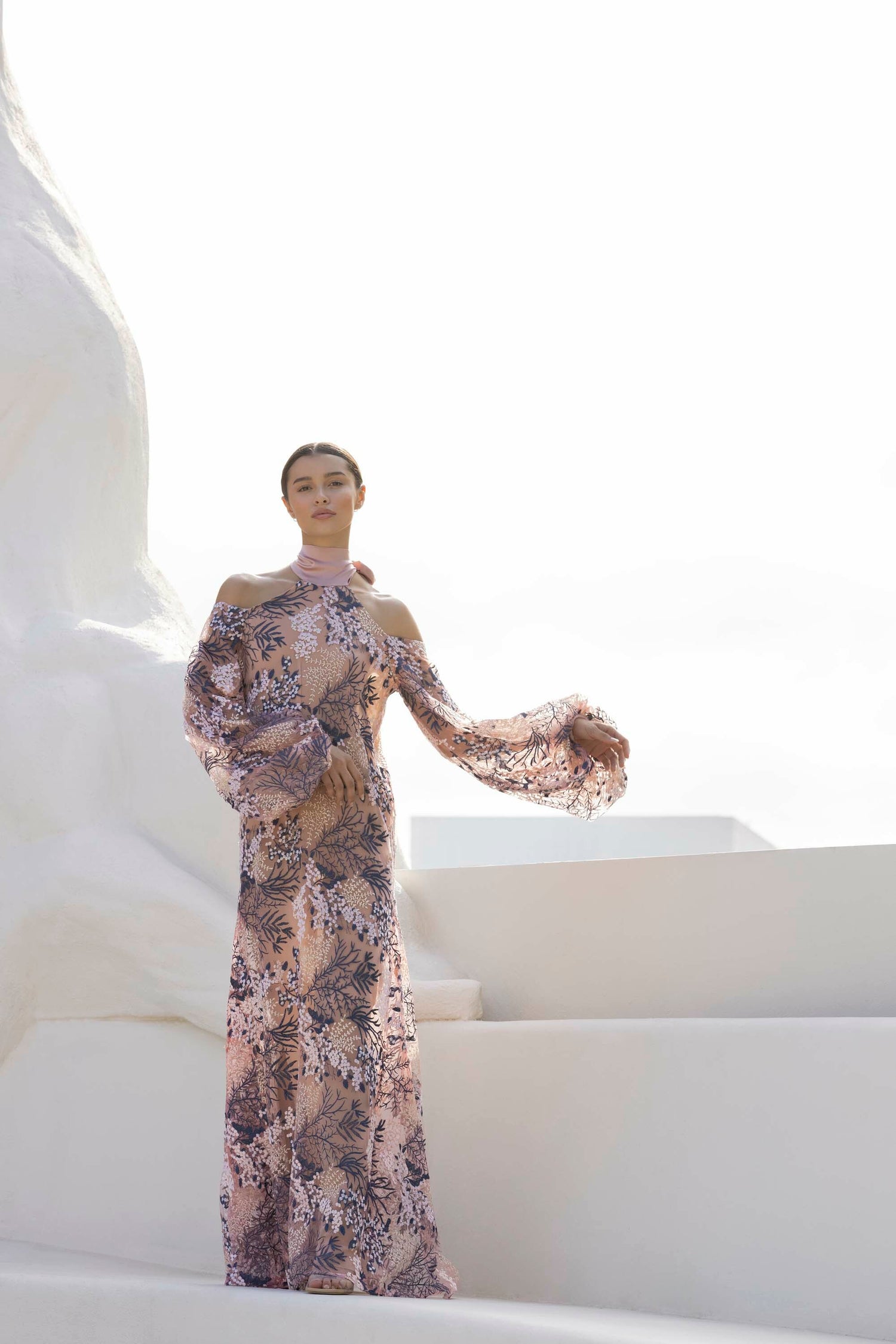 A person stands on white steps wearing an off-shoulder, long-sleeve floral dress. The background is bright and minimal.