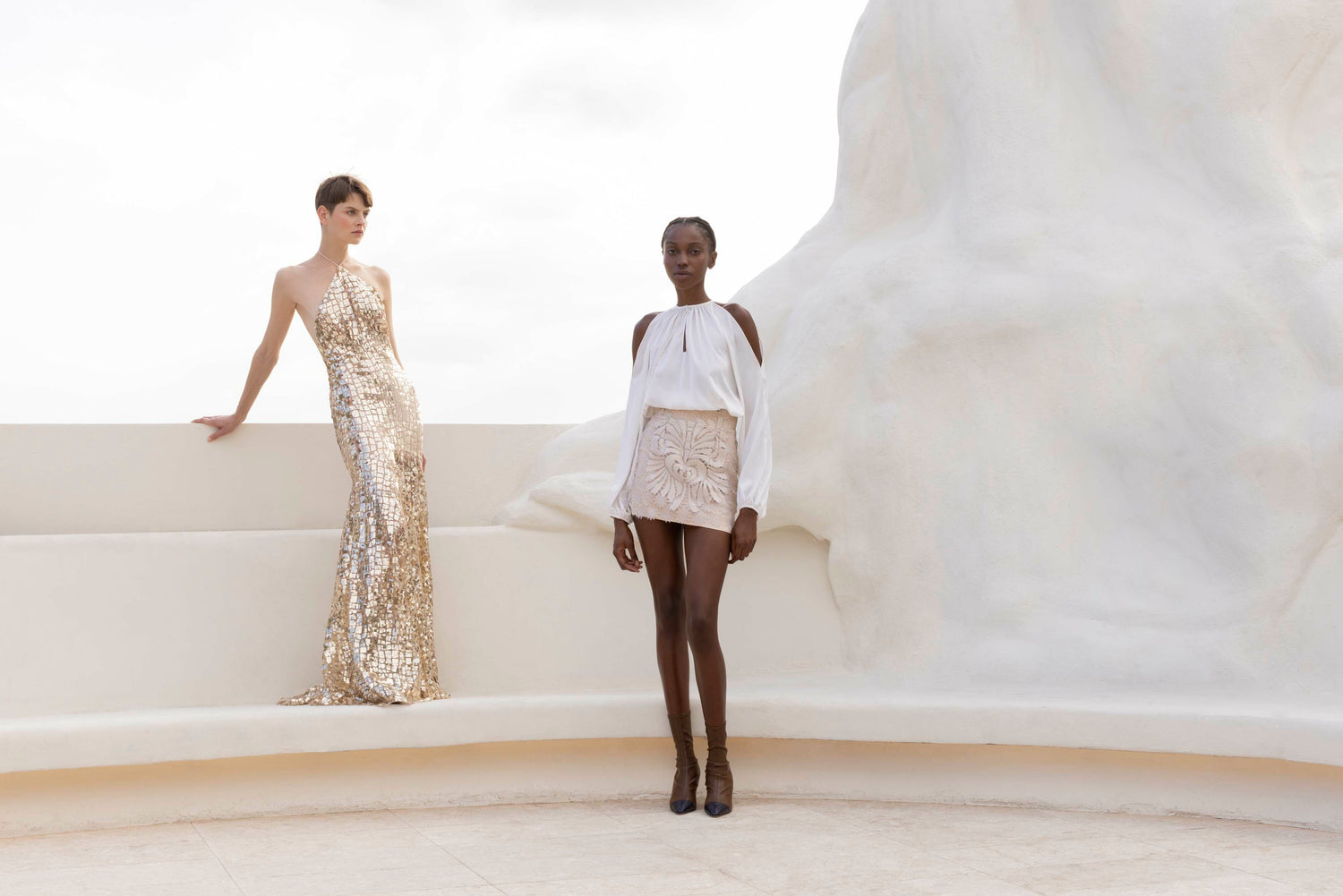 Two models pose in elegant outfits against a minimalist white architectural background.