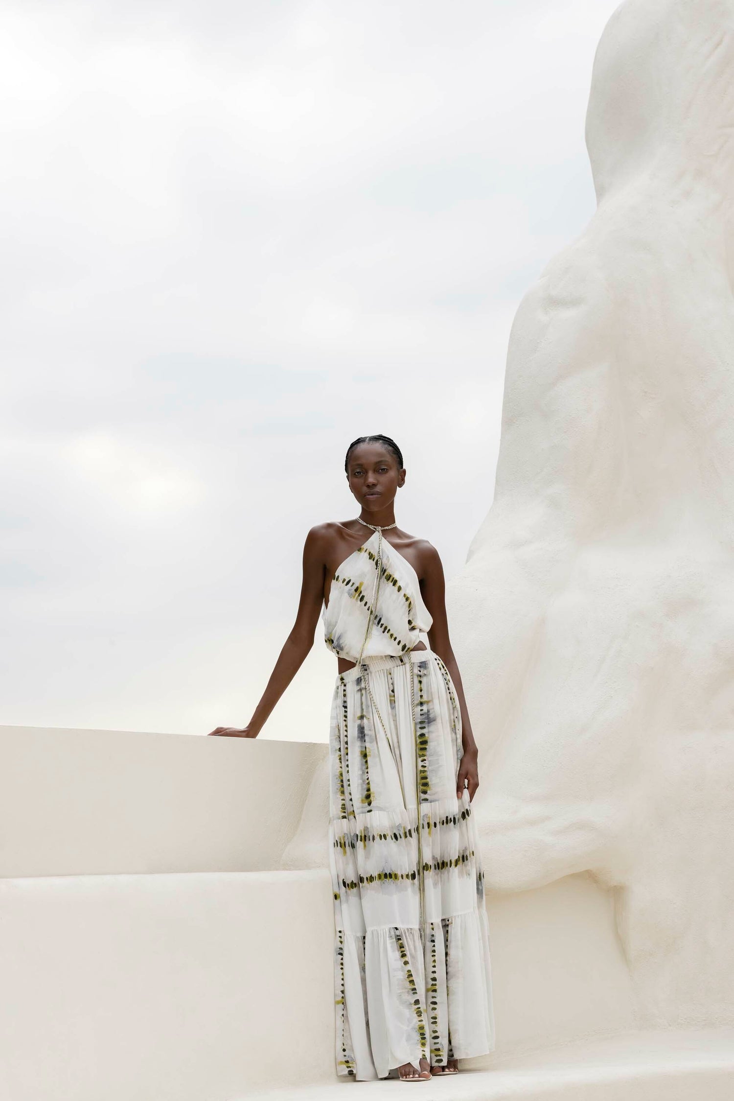 Person in a flowing white and yellow patterned dress stands outdoors against a light-colored sculptural background. Cloudy sky in the background.