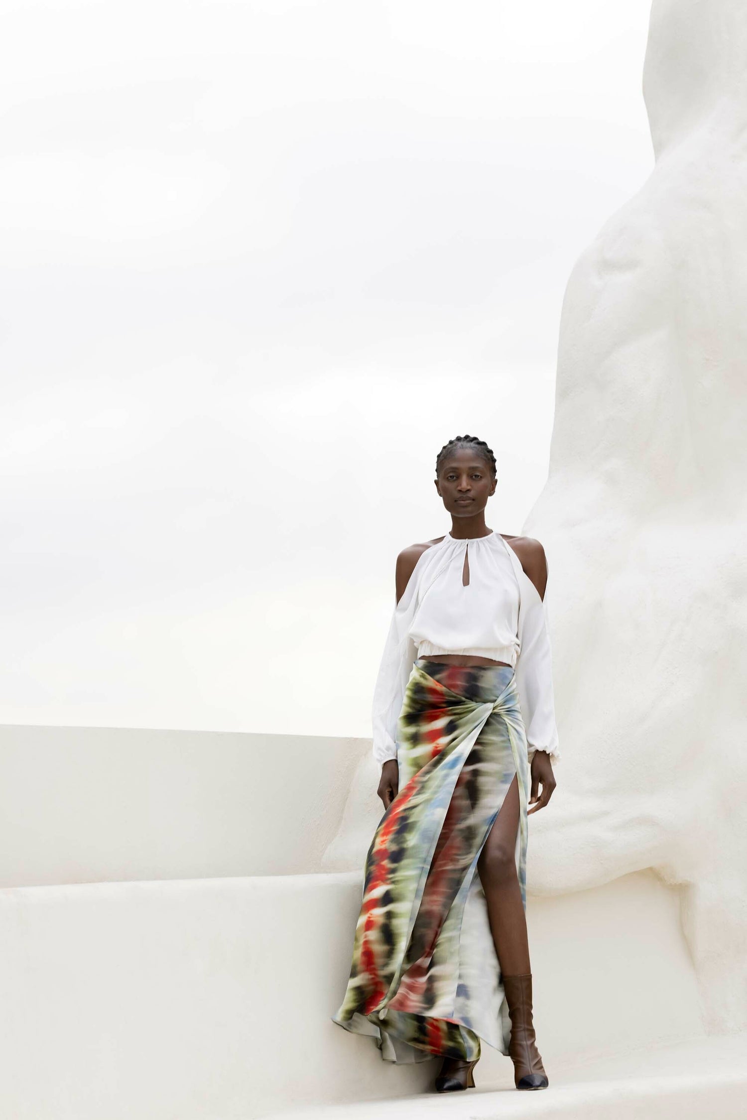 A woman stands against a white, abstract backdrop wearing a white blouse and a colorful skirt with a high slit.