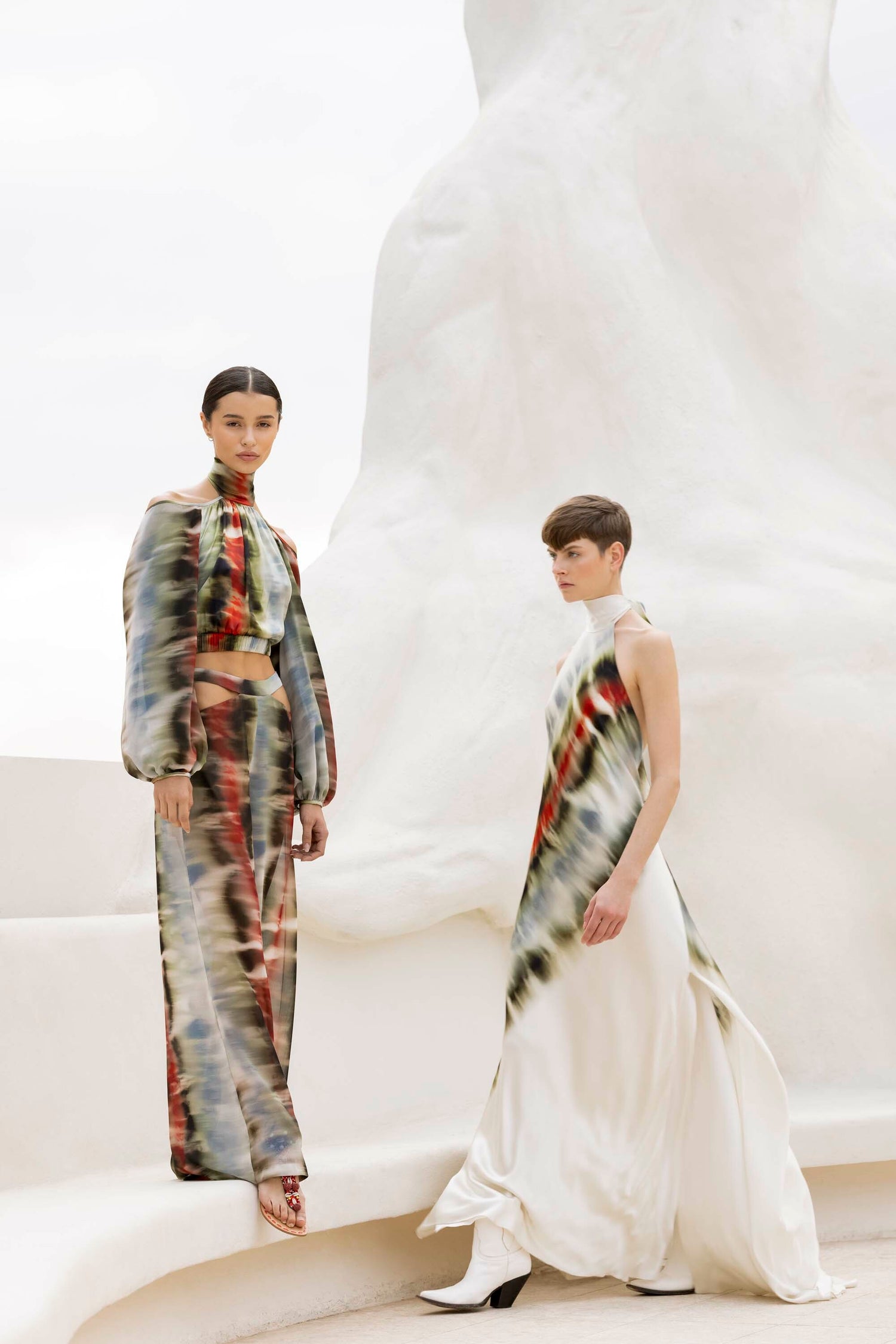 Two models wearing patterned dresses stand on pale, abstract architectural steps with large abstract forms in the background.