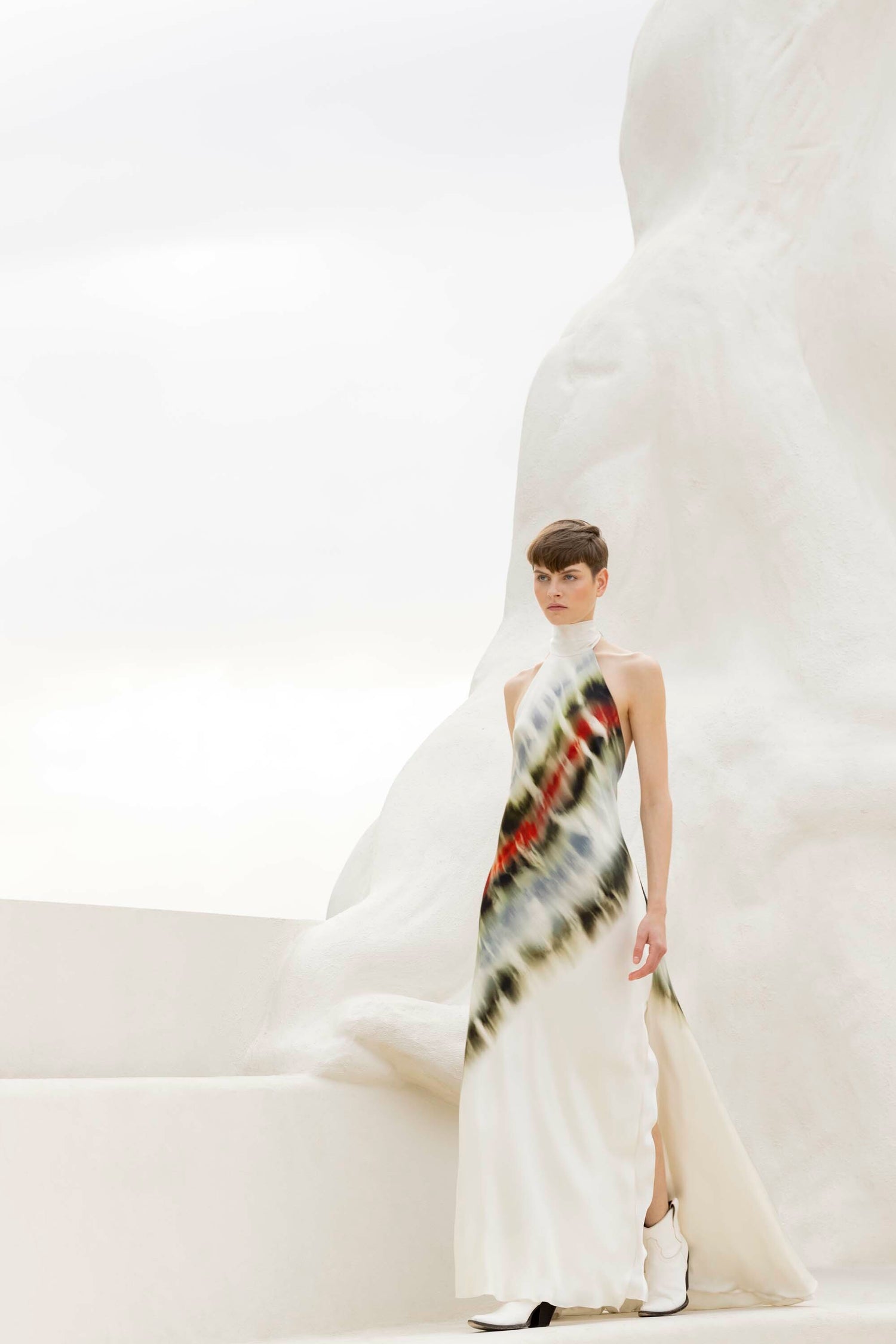 A person stands on white steps wearing a white dress with a colorful, diagonal pattern. The background features a smooth, abstract white sculpture.