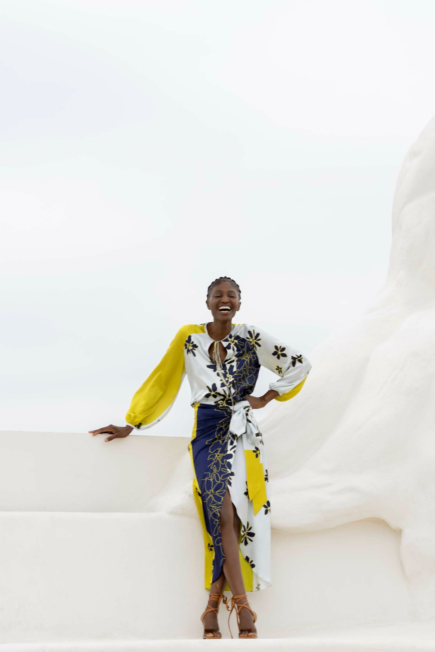 A person in a yellow and white floral dress stands outdoors, leaning on a white structure, smiling with one hand on their hip.