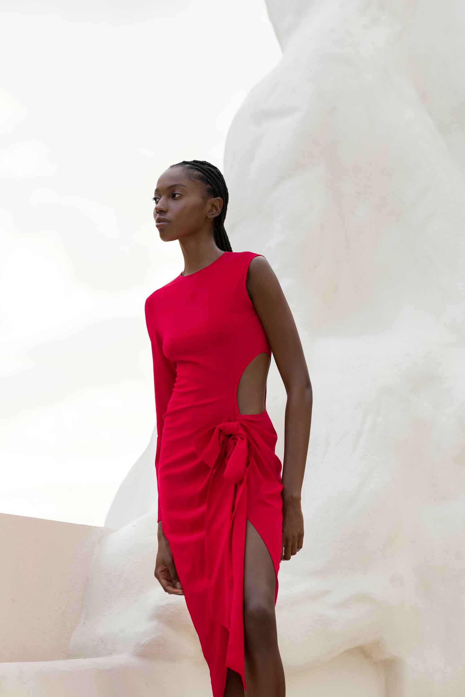 A person in a red dress with a side cut-out stands outdoors, with a large white sculpture in the background.