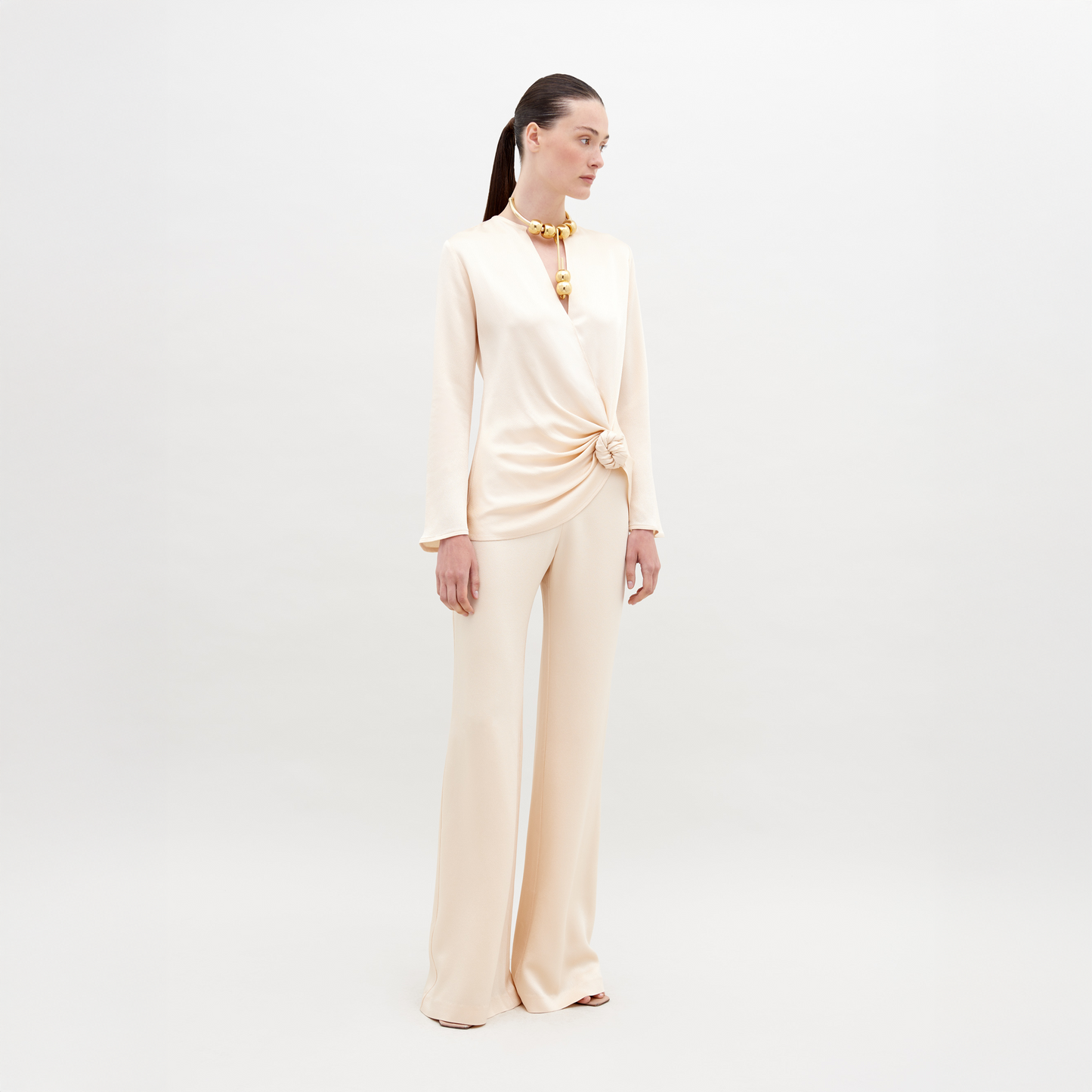 A woman stands against a plain background wearing an off-white, long-sleeve top with a knotted detail and matching Palermo Pant Vanilla. She has a high ponytail and is accessorized with a chunky necklace. Mark your calendars for November 15th, 2024—the ship by date for pre-orders of this chic ensemble.