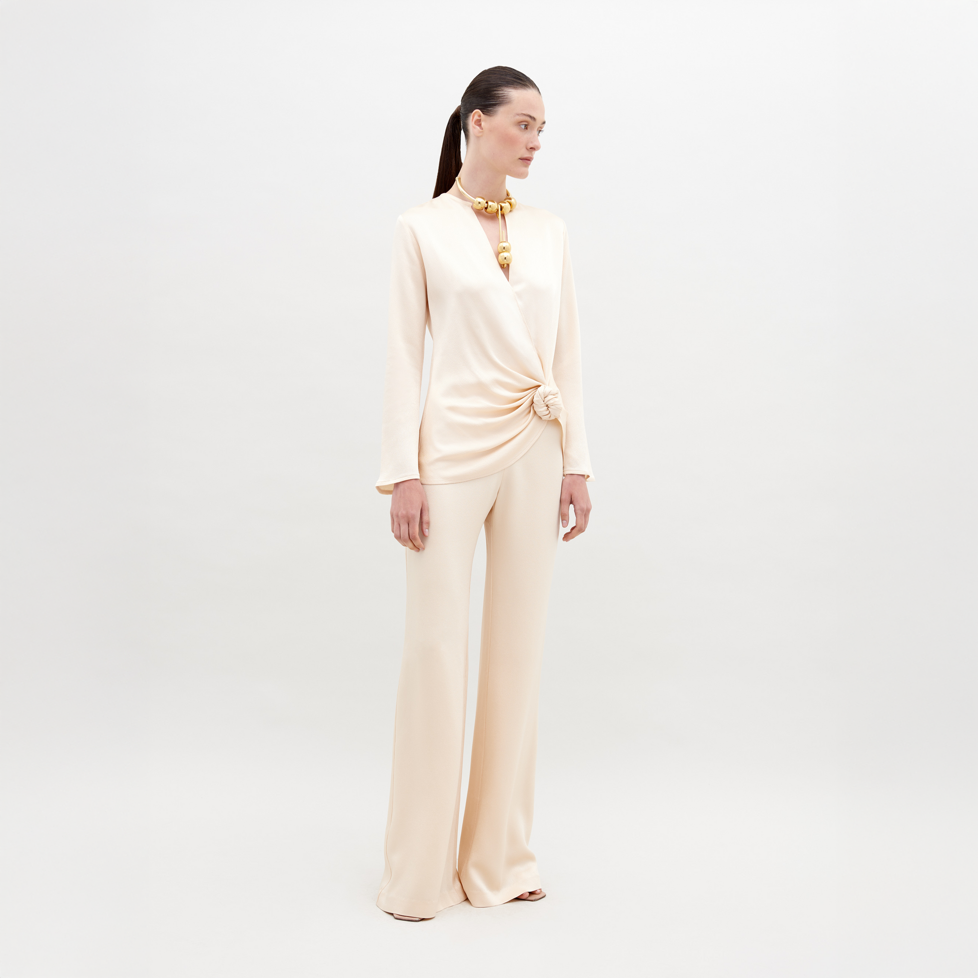 A woman stands against a plain background wearing an off-white, long-sleeve top with a knotted detail and matching Palermo Pant Vanilla. She has a high ponytail and is accessorized with a chunky necklace. Mark your calendars for November 15th, 2024—the ship by date for pre-orders of this chic ensemble.