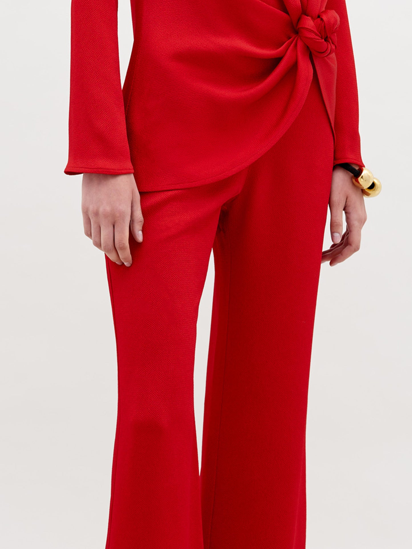 A person wears a long-sleeved, bright red top with a knotted waist and the matching high-waisted, wide-leg Palermo Pant. Their hand rests by their side, accessorized with a chunky gold bracelet.