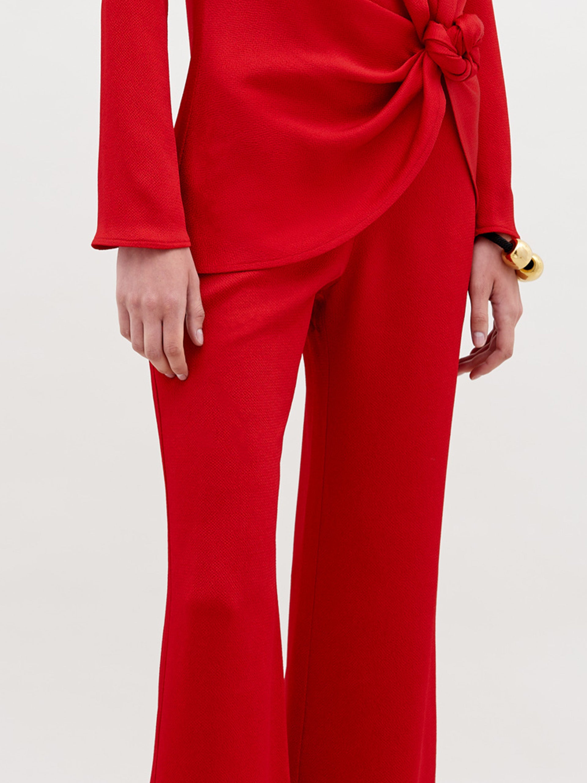 A person wears a long-sleeved, bright red top with a knotted waist and the matching high-waisted, wide-leg Palermo Pant. Their hand rests by their side, accessorized with a chunky gold bracelet.