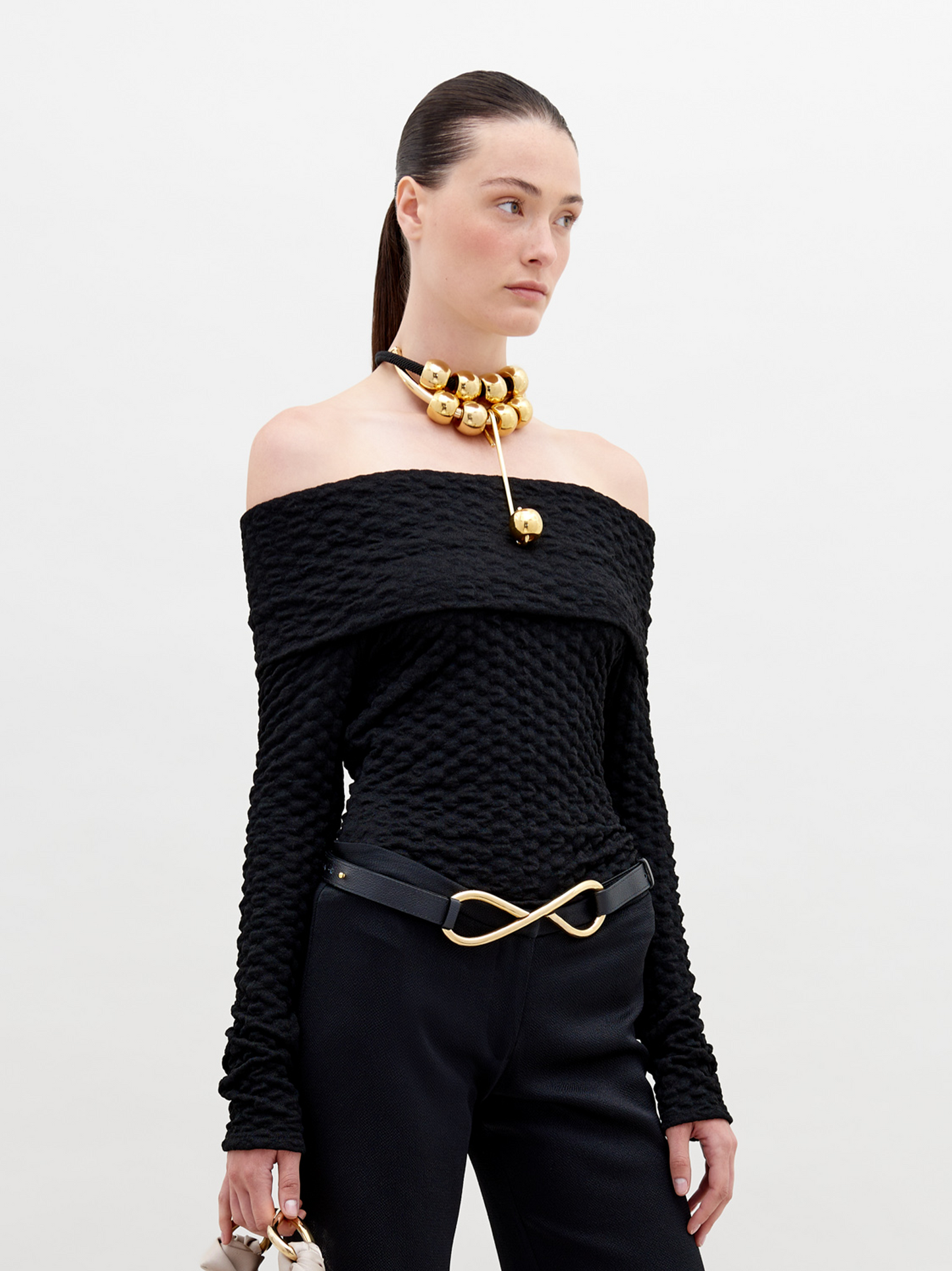 A woman is wearing an off-the-shoulder Palma Shirt Black, black pants with an infinity-shaped gold belt buckle, and a gold choker necklace with large round ornaments. She has her hair pulled back. Available for pre-order, this stylish ensemble will ship by November 15th, 2024.