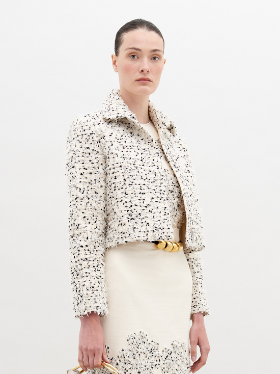 A woman with light skin and dark hair is wearing a Perla Jacket Beige Black Embroidered Flowers over a matching dress. Holding a small bag, she stands against a plain background. Pre-order now to ensure it ships by November 15th, 2024.