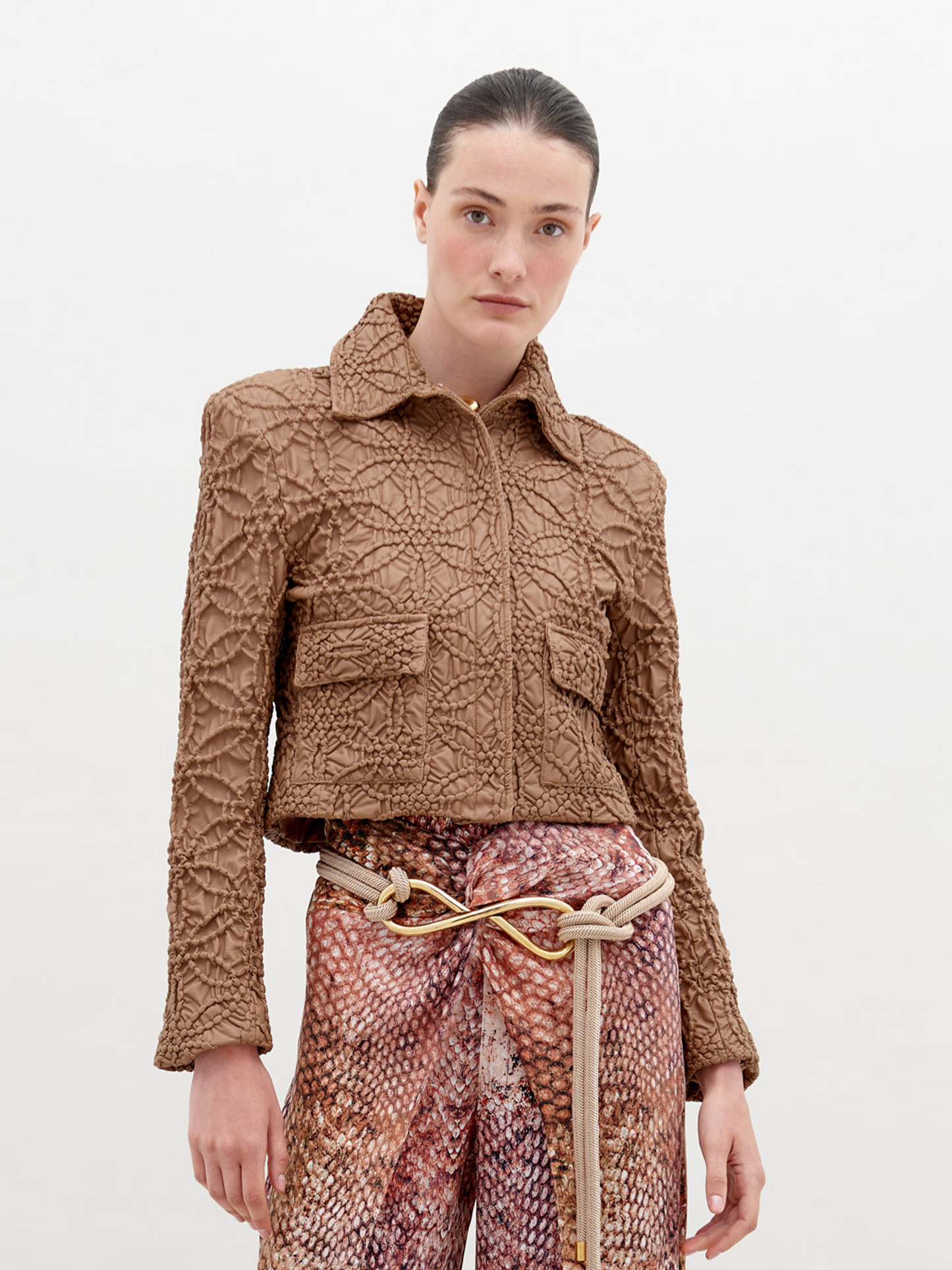 A woman stands against a plain background, wearing the Perla Jacket Brown and patterned high-waist pants. A decorative knot belt accentuates her waist. This stylish ensemble is available now, ensuring you can enhance your wardrobe immediately.