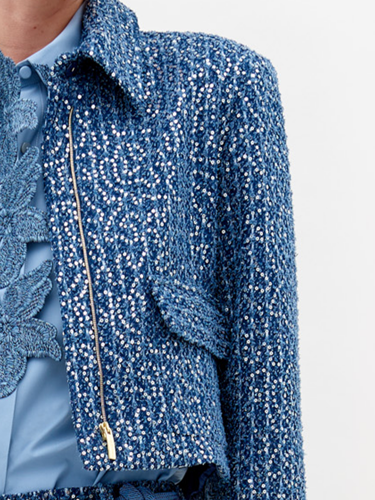 A person wearing a Perla Jacket Navy Sequins and pants, with a light blue shirt and a belt with pearl-like decorations, stands against a plain white background. Pre-order now to ensure delivery by November 15th, 2024.