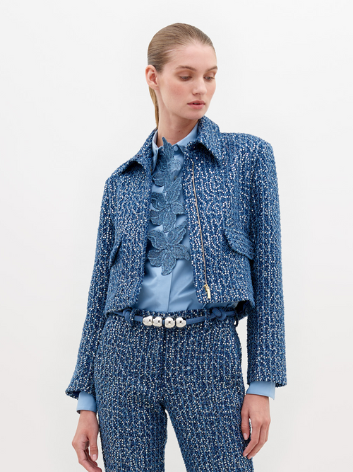 A person wearing a Perla Jacket Navy Sequins and pants, with a light blue shirt and a belt with pearl-like decorations, stands against a plain white background. Pre-order now to ensure delivery by November 15th, 2024.