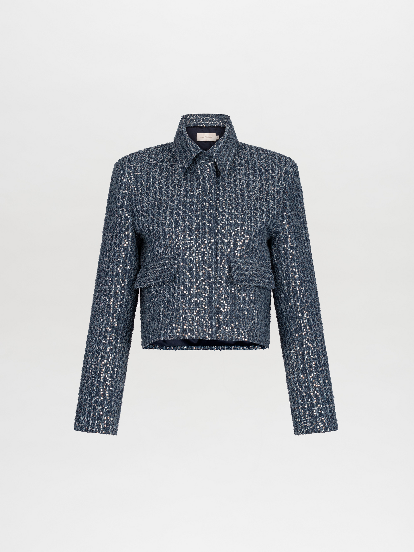 A person wearing a Perla Jacket Navy Sequins and pants, with a light blue shirt and a belt with pearl-like decorations, stands against a plain white background. Pre-order now to ensure delivery by November 15th, 2024.