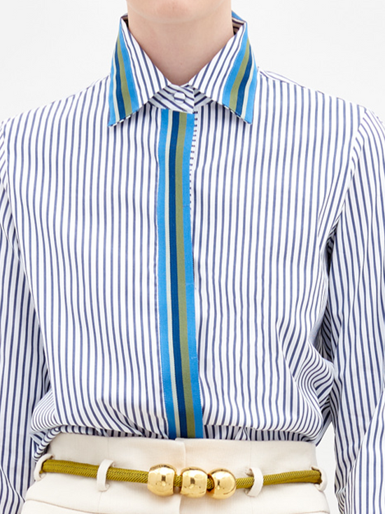 A person wearing a Premium Shirt White Blue Stripes with a blue accent, white pants, and a yellow belt holds a small bag in their hand. The background is plain white. This stylish ensemble will be available for pre-order starting November 15th, 2024.