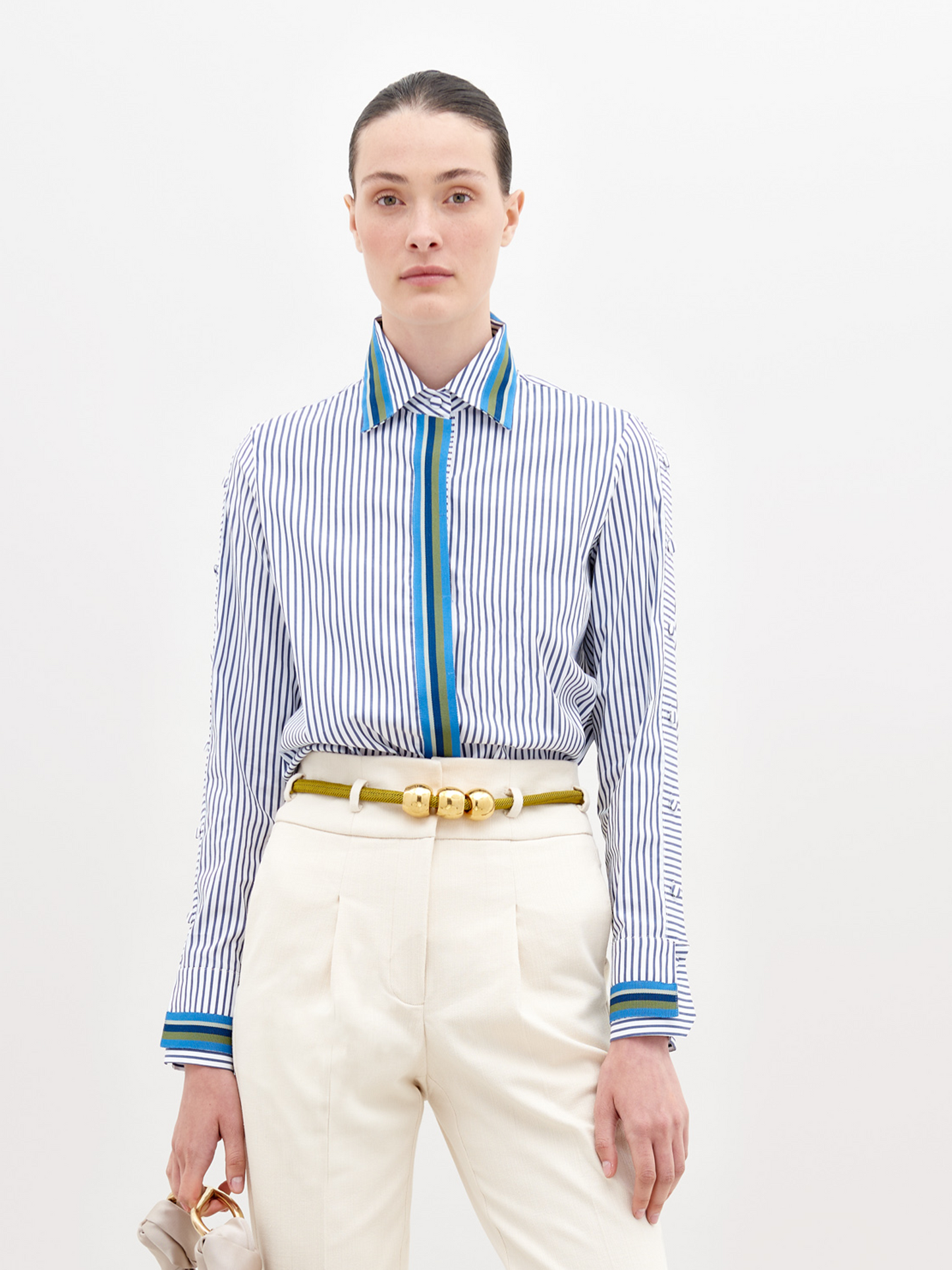 A person wearing a Premium Shirt White Blue Stripes with a blue accent, white pants, and a yellow belt holds a small bag in their hand. The background is plain white. This stylish ensemble will be available for pre-order starting November 15th, 2024.