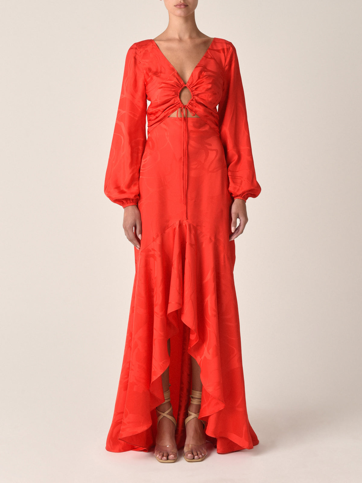 A woman wears the Charlize Dress Crimson Jacquard, featuring long sleeves, a V-neck with front cutout, keyhole detail, high-low asymmetrical skirt, and a subtle pattern. She pairs it with strappy sandals.