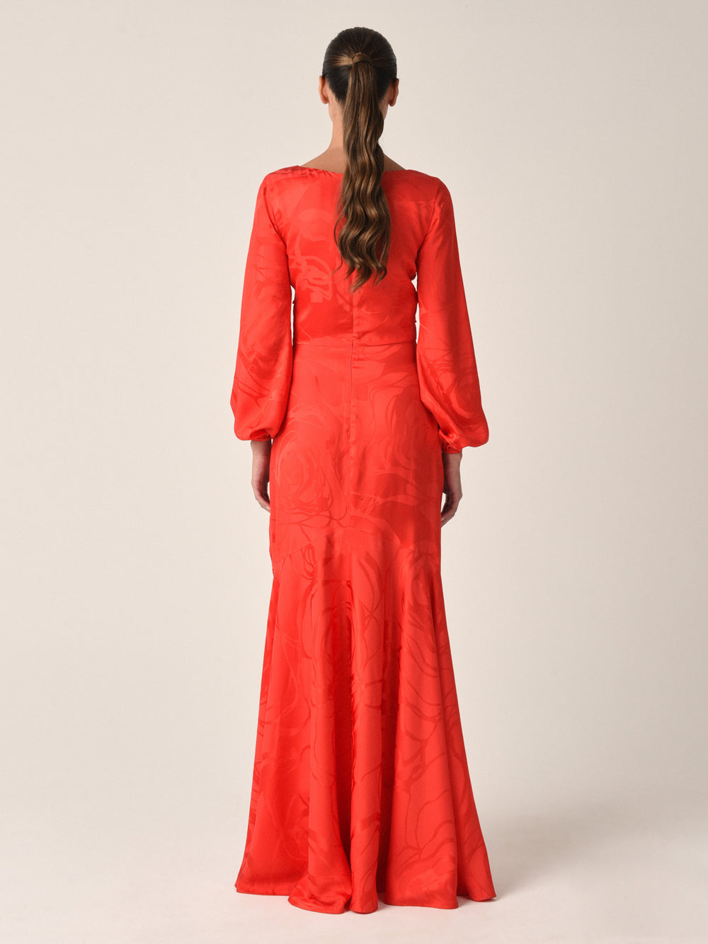 A woman with brown hair in a loose ponytail faces away, wearing the Charlize Dress Crimson Jacquard—featuring subtle floral patterns, long puffed sleeves, and a fitted waist—against a plain light-colored background.