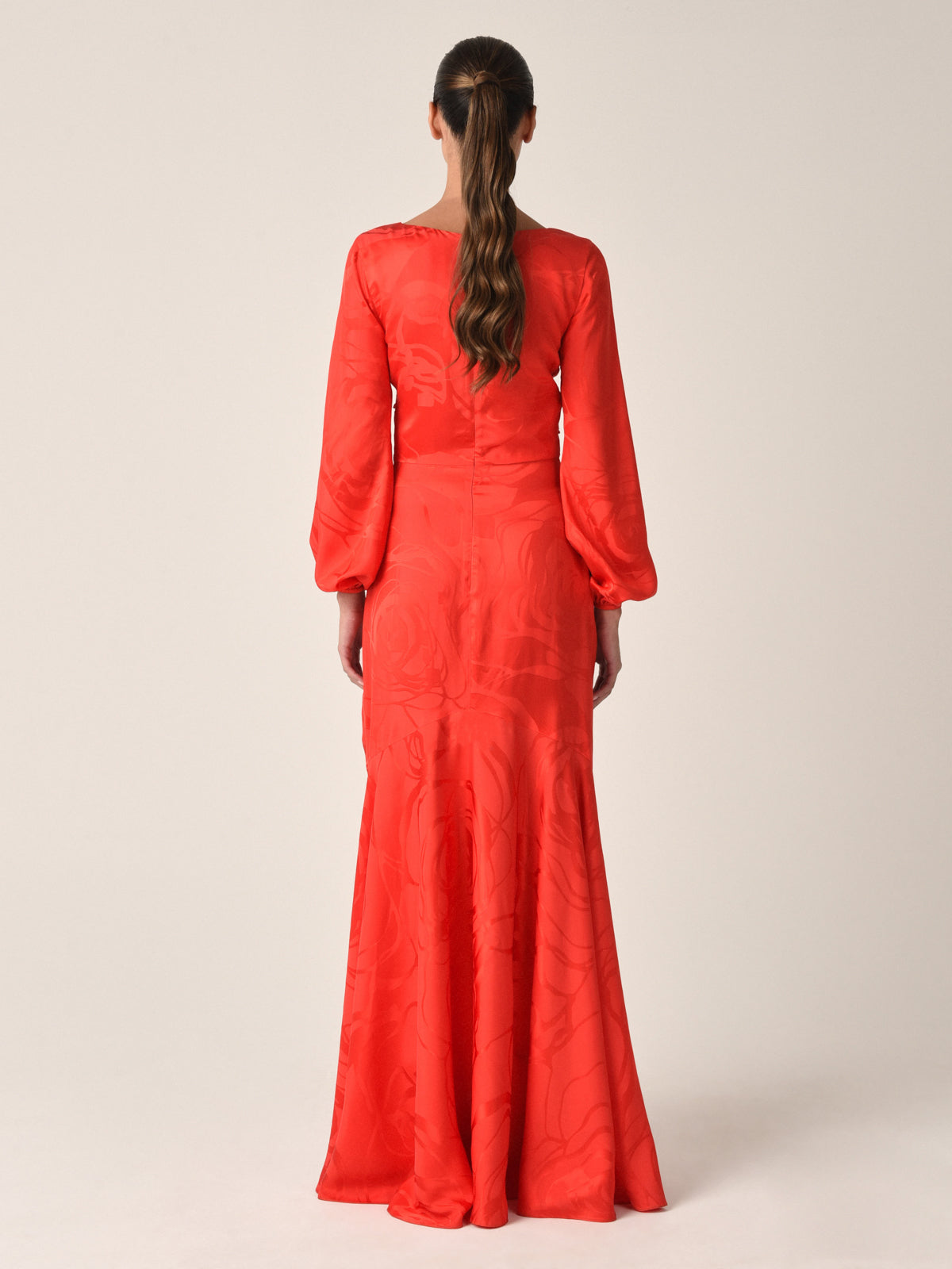A woman with brown hair in a loose ponytail faces away, wearing the Charlize Dress Crimson Jacquard—featuring subtle floral patterns, long puffed sleeves, and a fitted waist—against a plain light-colored background.