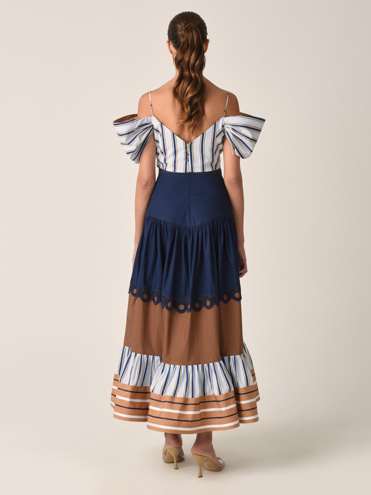 The Justine Skirt Camel Navy Stripes is a long, tiered midi skirt featuring a navy blue top section, a brown middle section adorned with cotton guipure lace detail, and a striped bottom section in shades of blue, brown, and white.