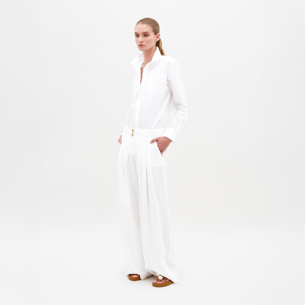 A person stands against a plain background, wearing an all-white outfit featuring the Opal Pant White—a pair of wide-legged pants—combined with a long-sleeve shirt with a high collar, their hands casually in their pockets. Pre-order now to secure your style for February 15th, 2025.