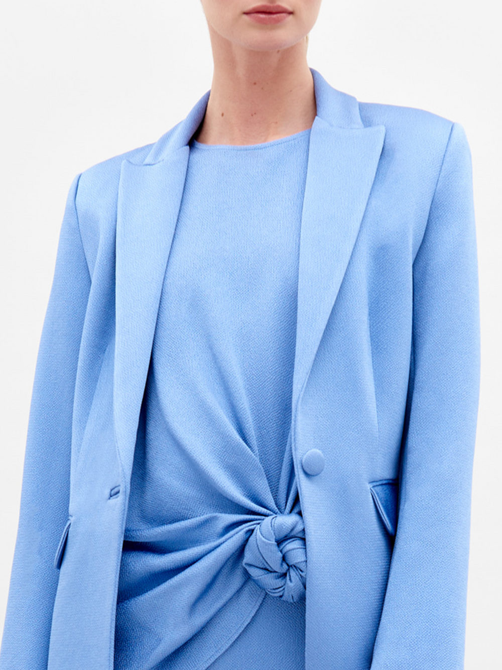 A woman in a light blue Rebeca Jacket with hammered satin, single-button closure, and matching knotted top and pants holds sunglasses against a plain white background.