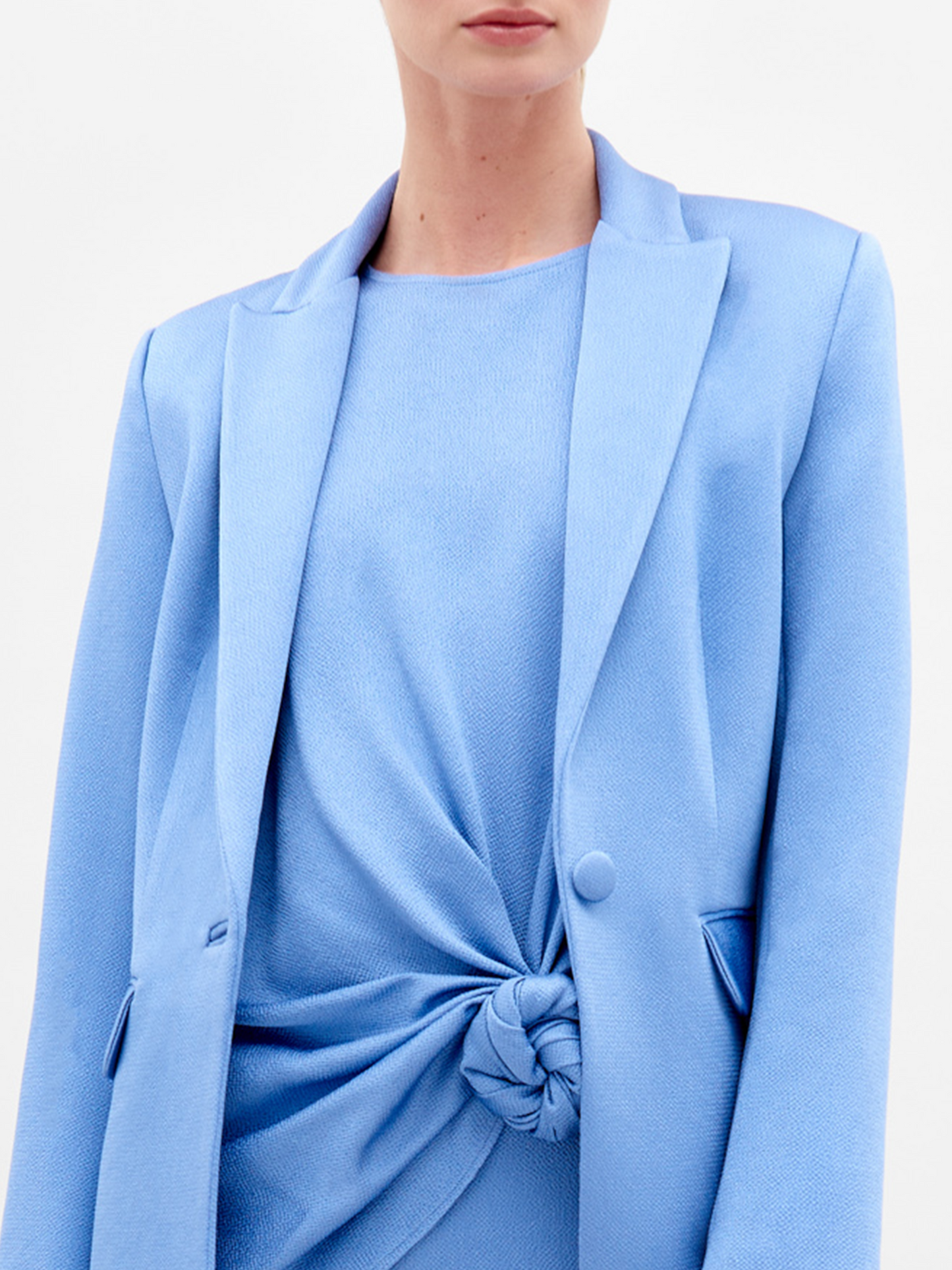 A woman in a light blue Rebeca Jacket with hammered satin, single-button closure, and matching knotted top and pants holds sunglasses against a plain white background.