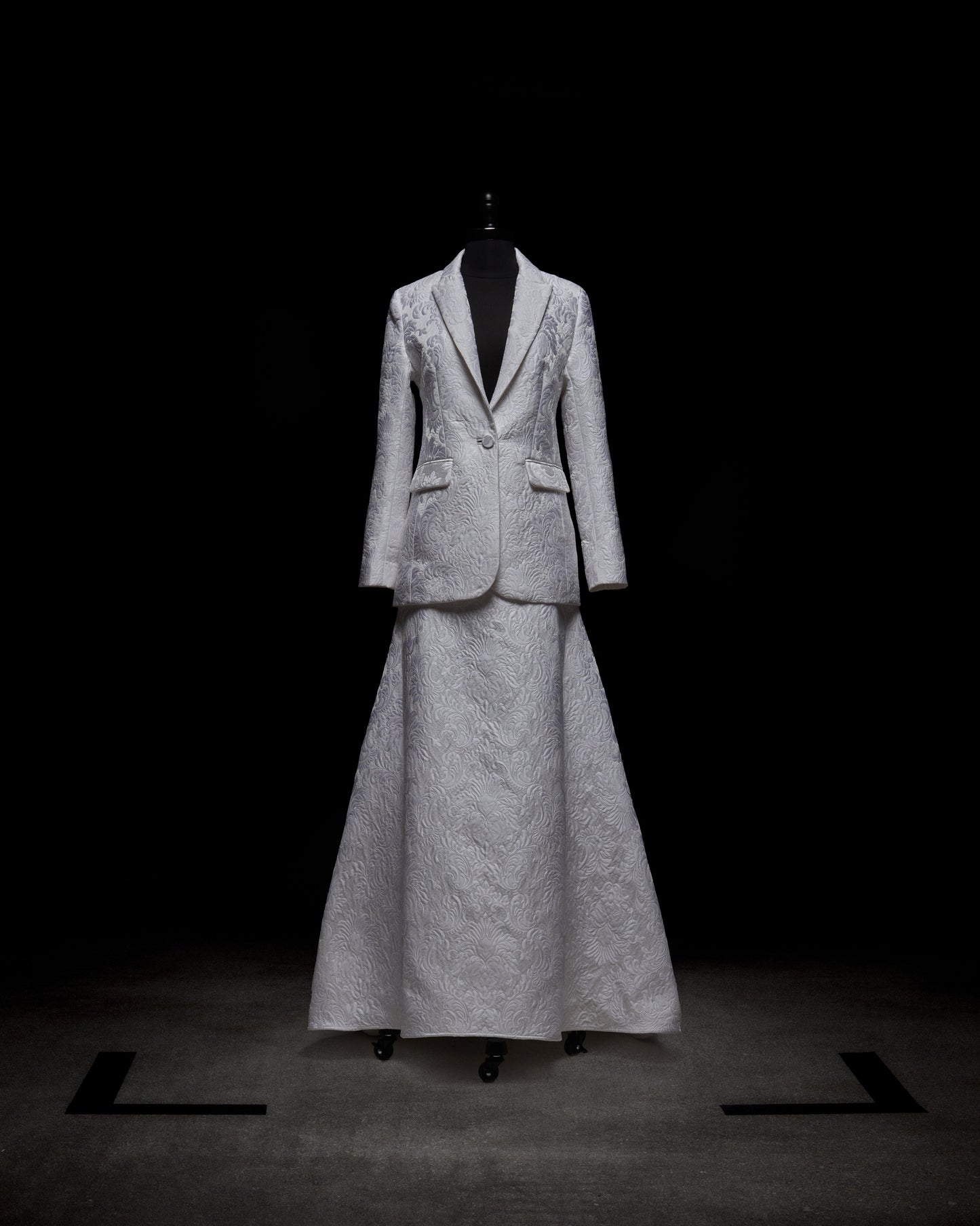 The Imola Skirt, a high-waisted maxi in a light, textured fabric, is shown with a matching suit jacket on a mannequin set against a dark background, with soft lighting accentuating the garment’s refined details.
