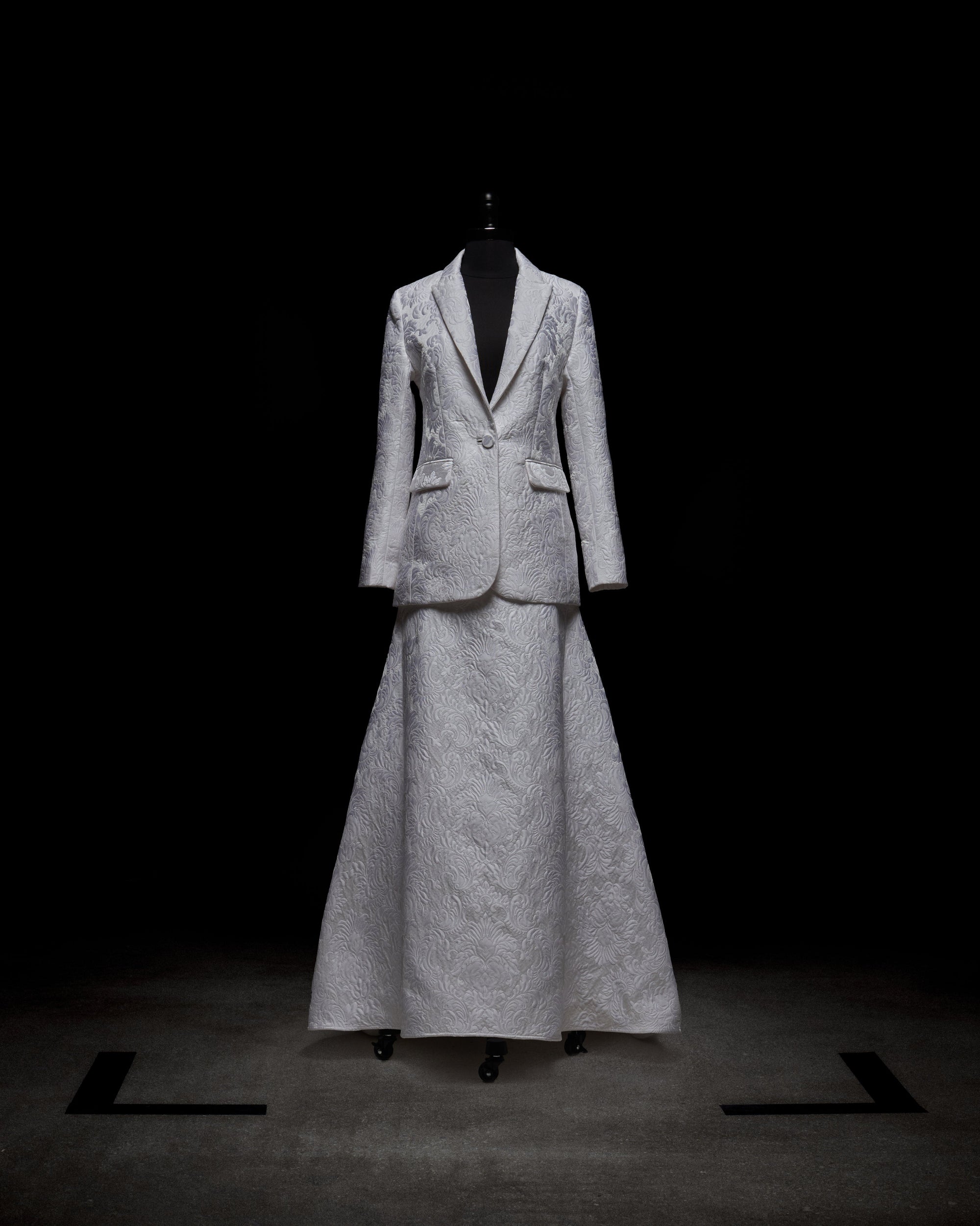 The Imola Skirt, a high-waisted maxi in a light, textured fabric, is shown with a matching suit jacket on a mannequin set against a dark background, with soft lighting accentuating the garment’s refined details.